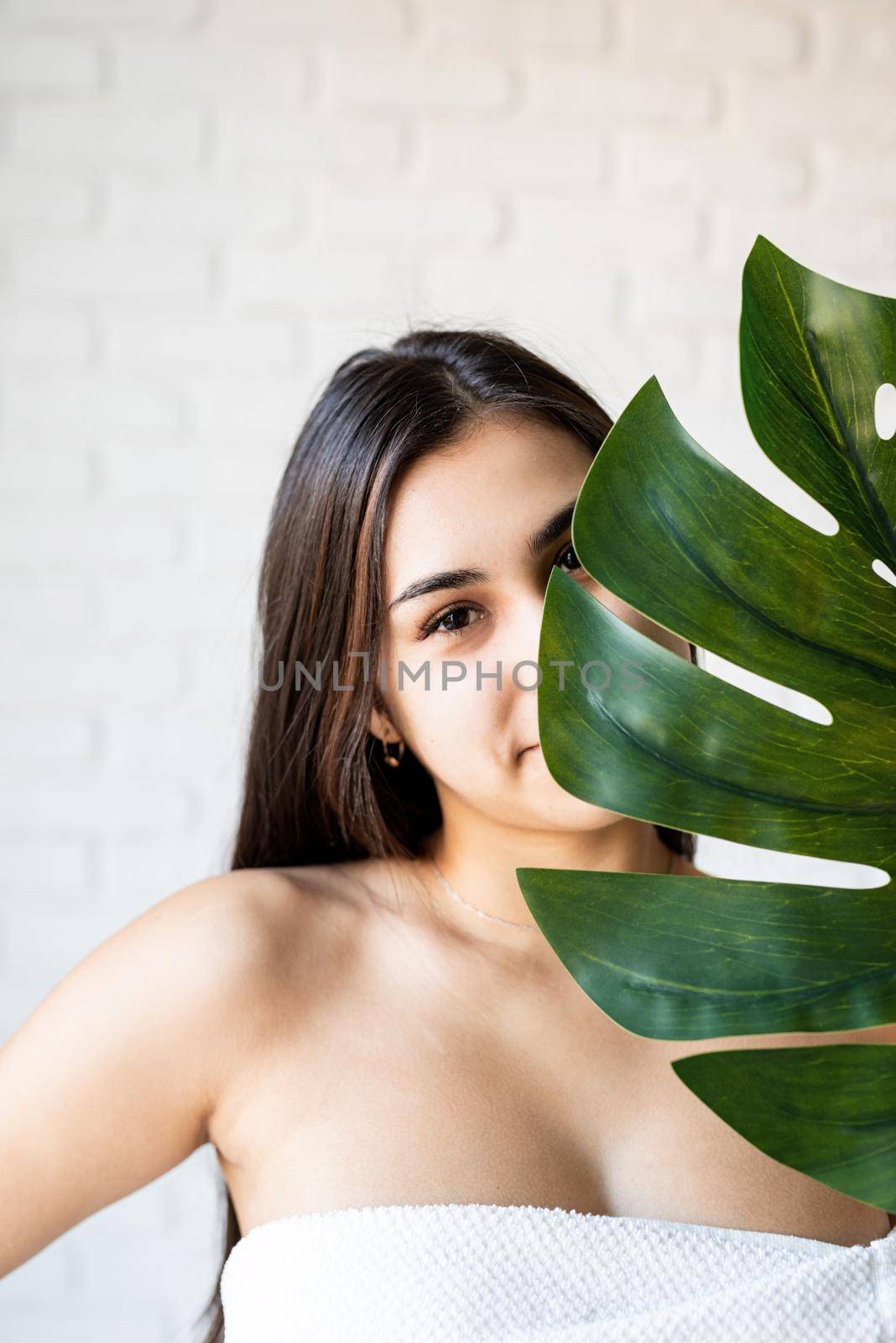 Spa Facial Mask. Spa and beauty. Happy beautiful brunette woman wearing bath towels holding a green monstera leaf in front of her face