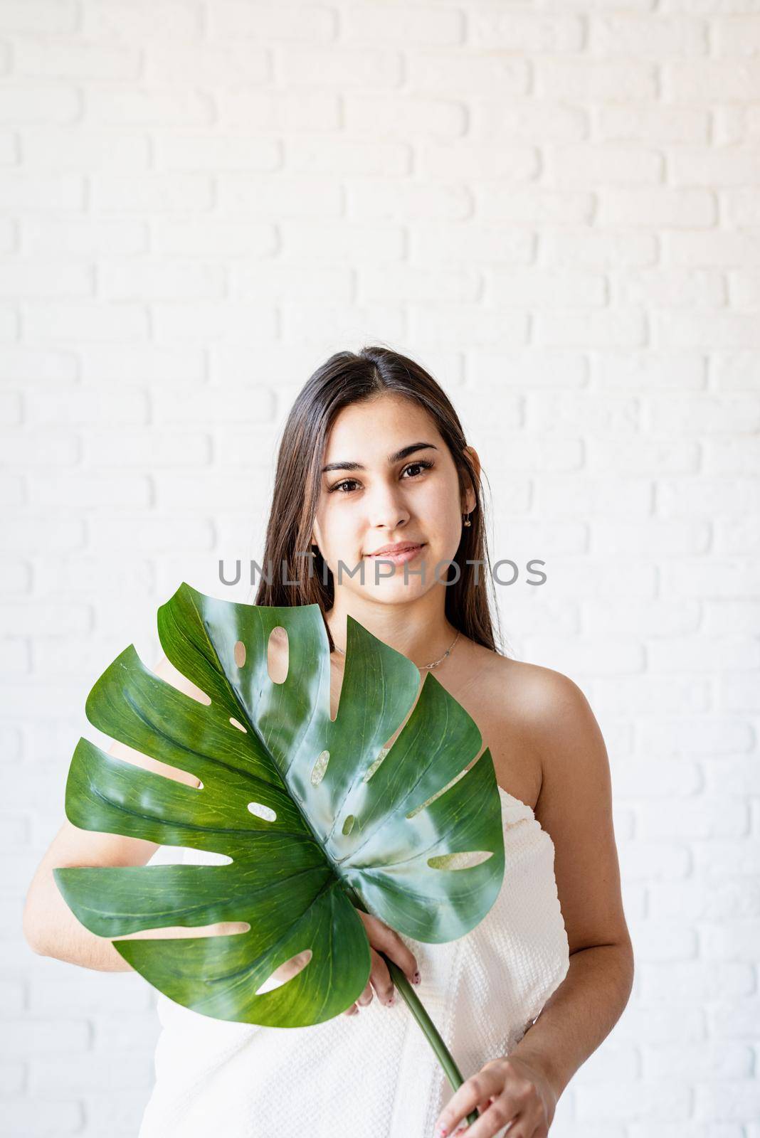 Spa Facial Mask. Spa and beauty. Happy beautiful brunette woman wearing bath towels holding a green monstera leaf in front of her face
