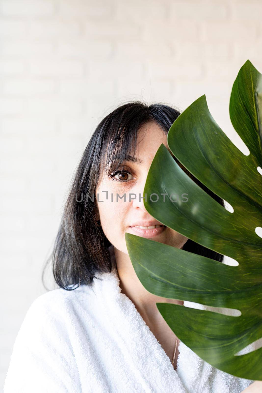 Spa Facial Mask. Spa and beauty. Happy beautiful brunette middle eastern woman wearing bath robes holding a green monstera leaf in front of her face