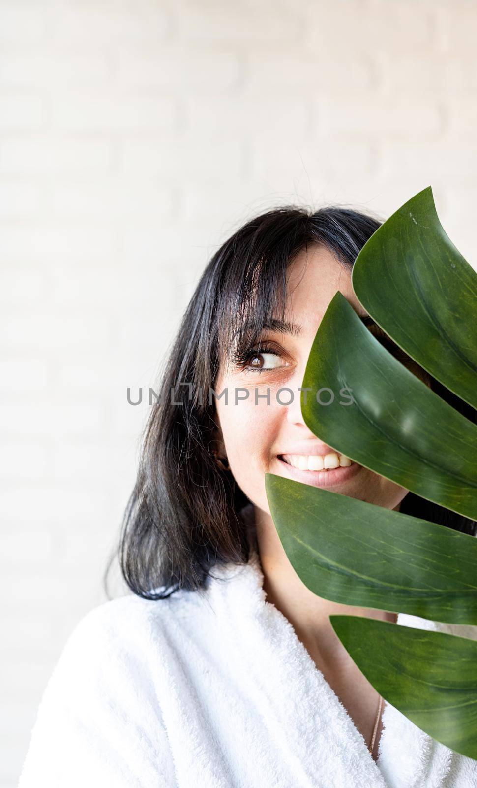 Spa Facial Mask. Spa and beauty. Happy beautiful brunette middle eastern woman wearing bath robes holding a green monstera leaf in front of her face