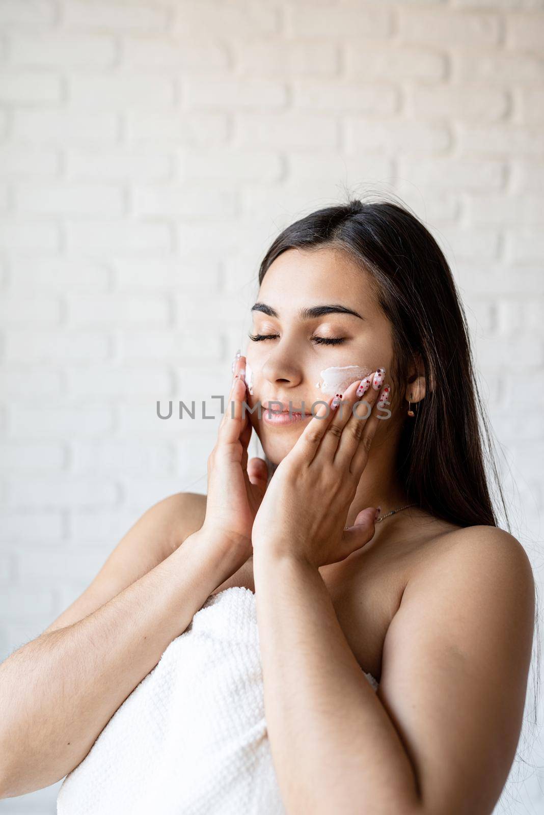 Happy beautiful woman wearing bath robes applying facial cream on her face by Desperada