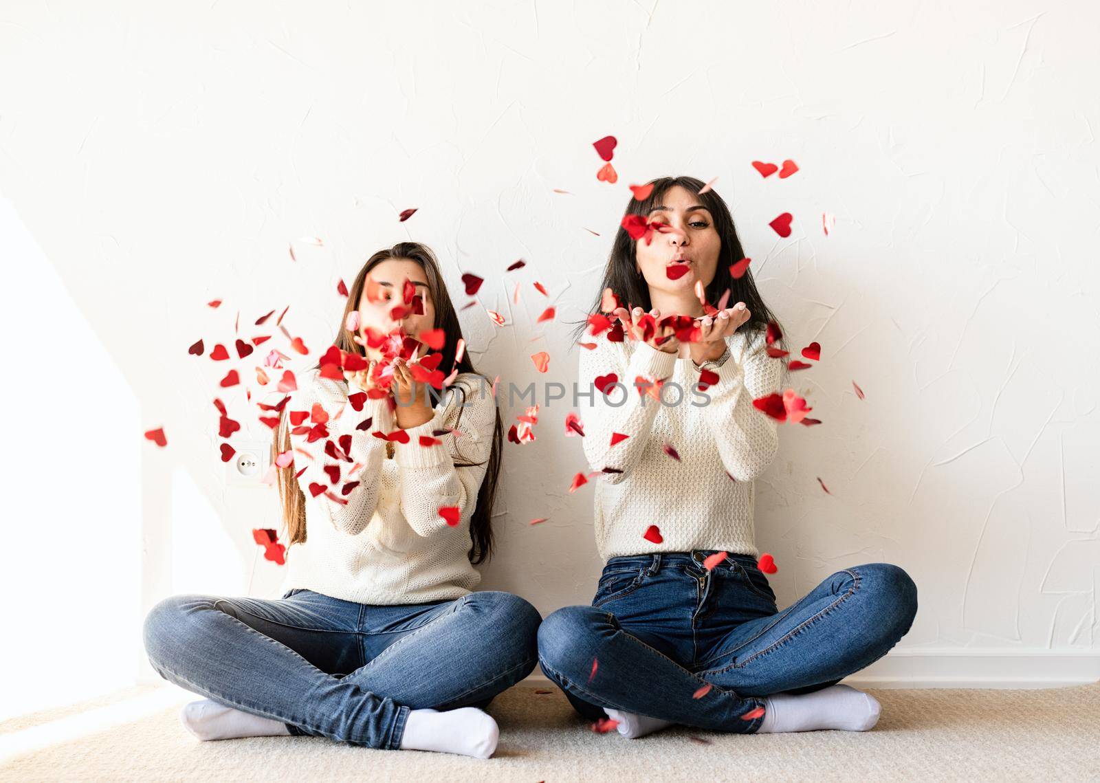 Two best friends having fun at home blowing red heart shape confetti