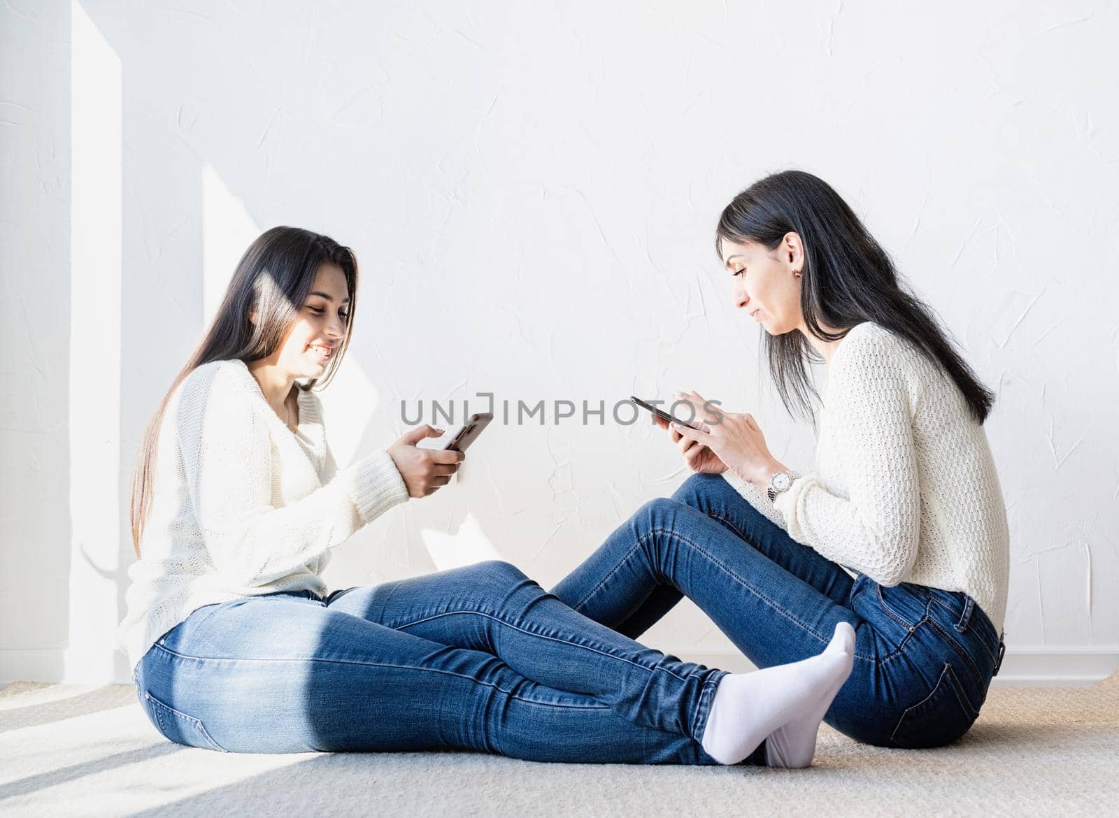 Two beautiful laughing women in jeans and white sweaters texting to friends using mobile phone