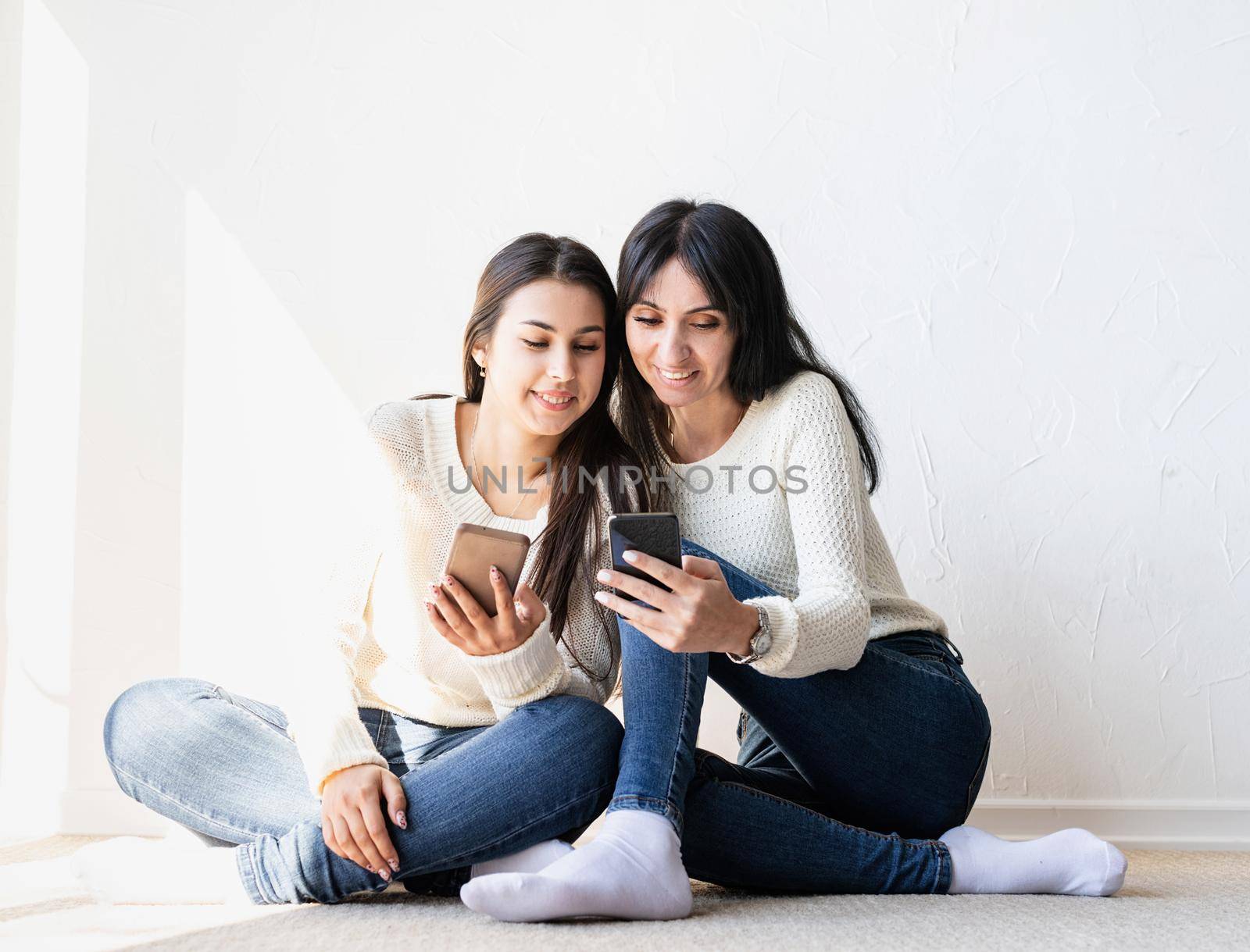 Two beautiful laughing women in jeans and white sweaters texting to friends using mobile phone