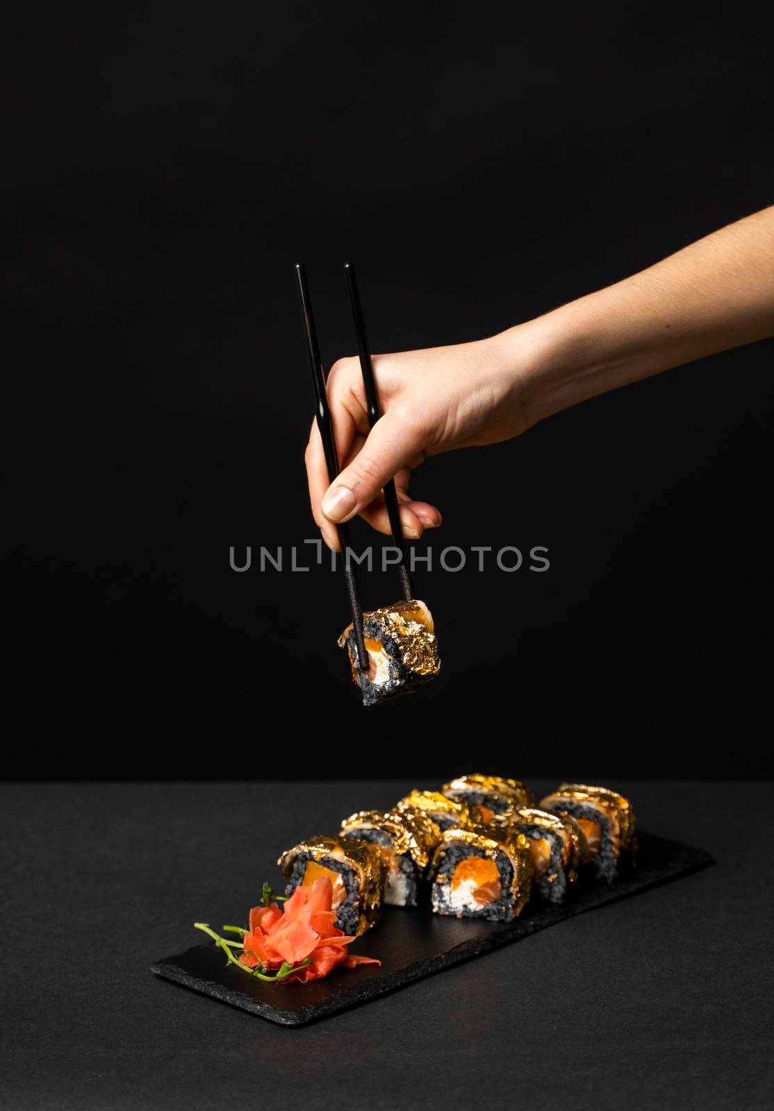 Hand with black chopsticks holds a sushi. Custom sushi roll with cuttlefish ink, salmon, cream cheese, pepper pumpkin, eel and edible gold leaf with ginger, wasabi on a square plate on black table