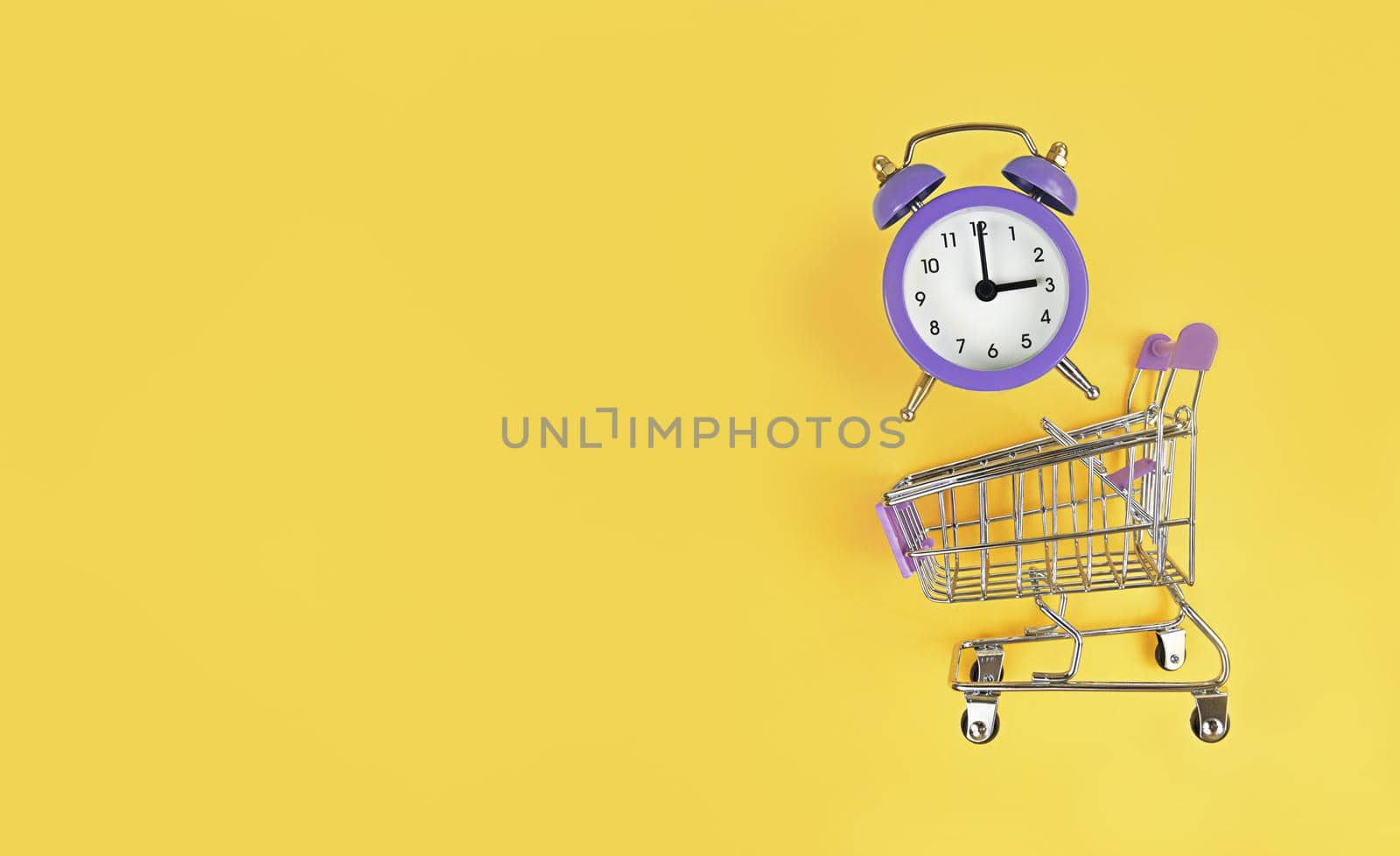 Supermarket trolley and violet alarm clock on a yellow background with copy space. Buy time, busy concept.