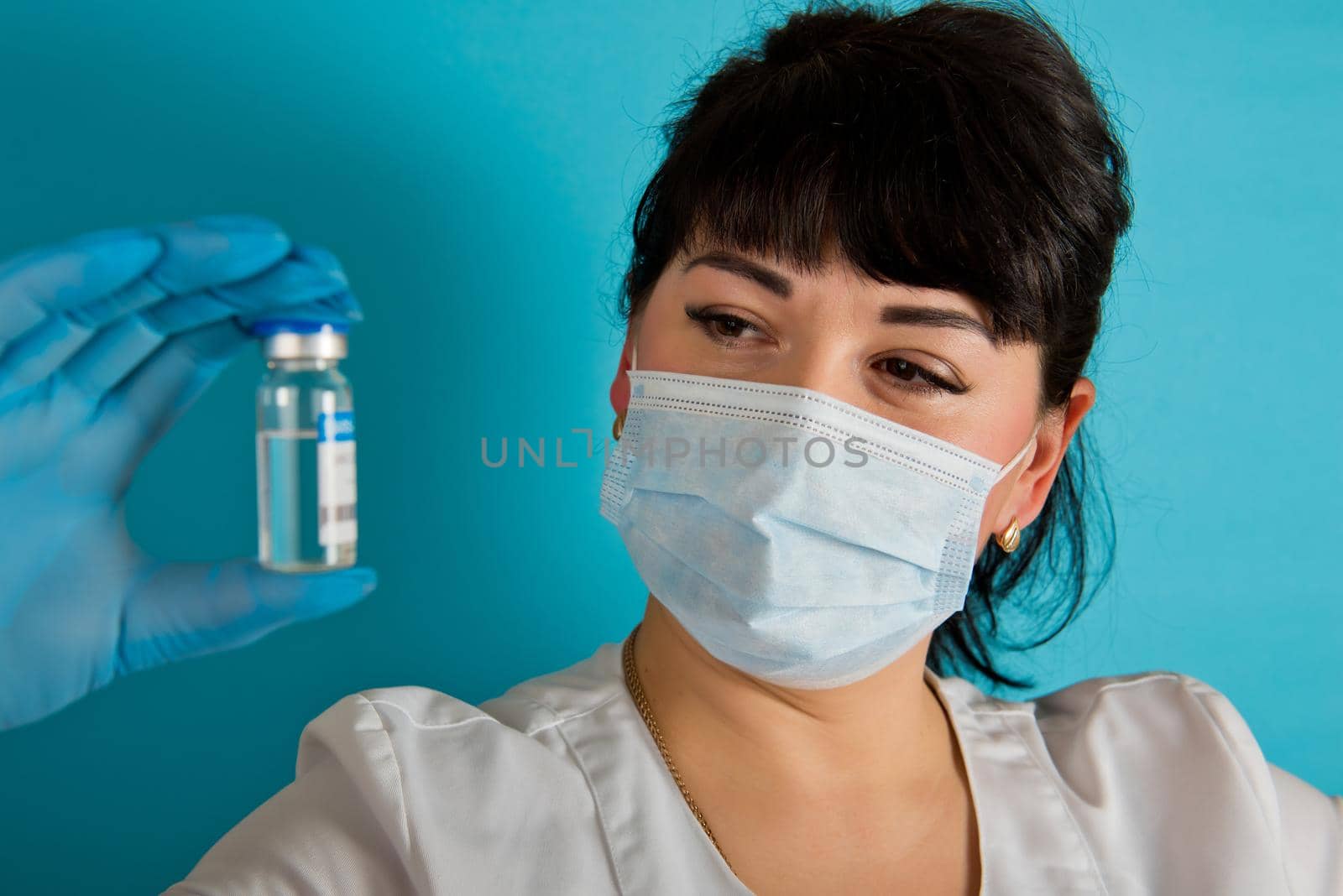 Young nurse in a medical mask holding vial of the Covid-19 coronavirus vaccine on a blue background. Close-up.