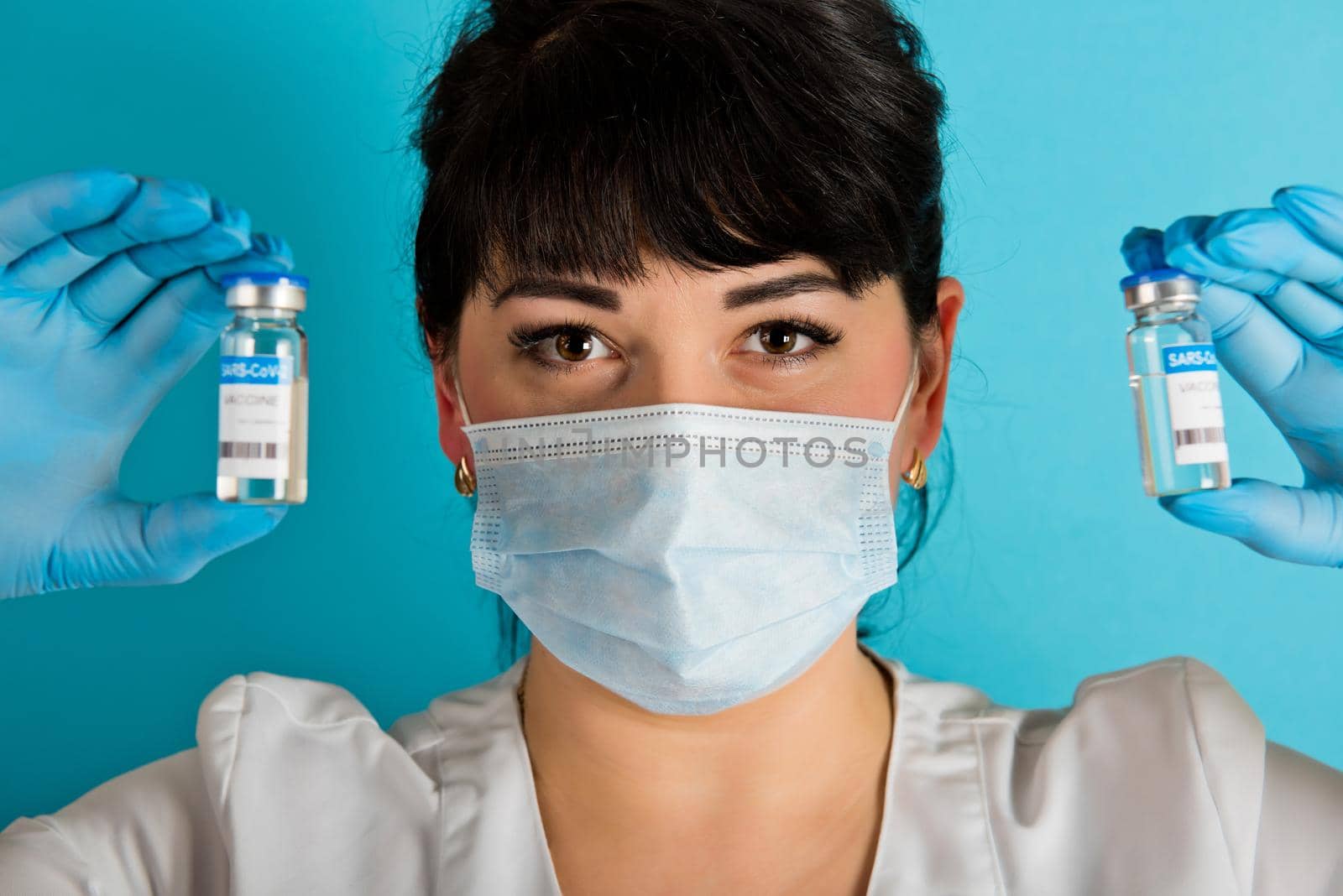 Young nurse in a medical mask holding two vials of the Covid-19 coronavirus vaccine on a blue background. Close-up.