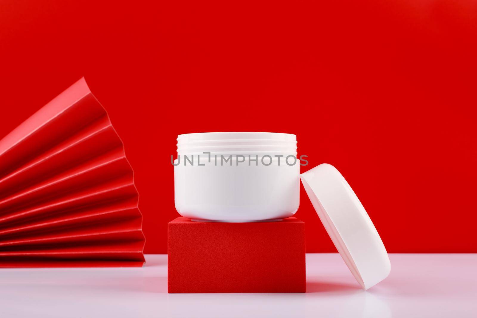 Selective focus, close up of opened jar with cream or mask on red pedestal against red background decorated with waver by Senorina_Irina