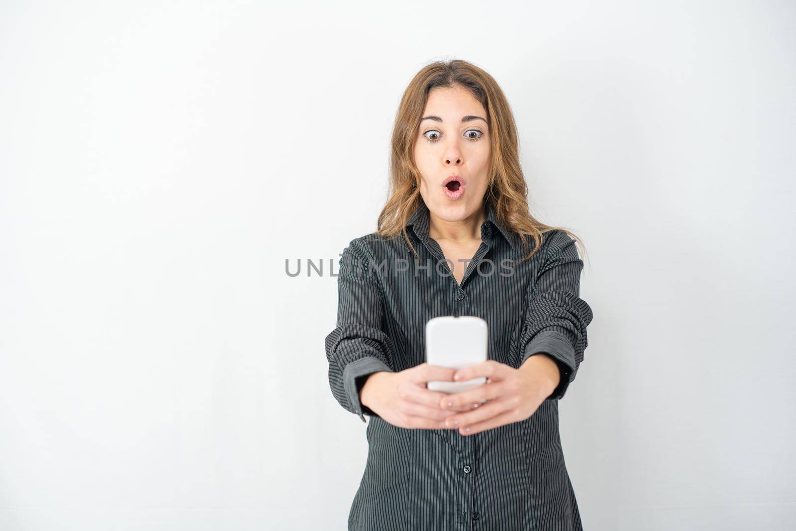 Surprised isolated beautiful young woman posing in study shot looking smartphone with eyes and mouth wide open. Young female technology addicted with wow winning expression on face