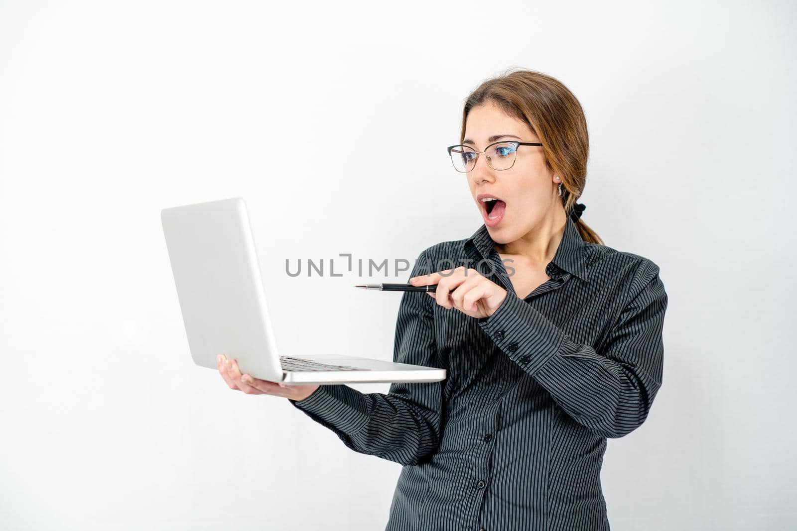 Surprised isolated beautiful young woman posing in study shot holding laptop in hand pointing with pen the screen wearing office business clothes. Caucasian female employee in wow expressions watch pc