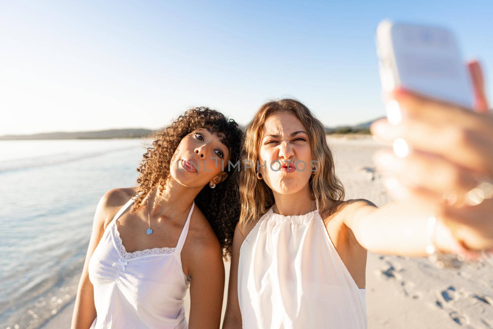 Beautiful mixed race female love couple making faces doing self portrait on beach - Two lesbian pretty women having fun using smartphone to sharing diversity - Selective focus on right girl face by robbyfontanesi