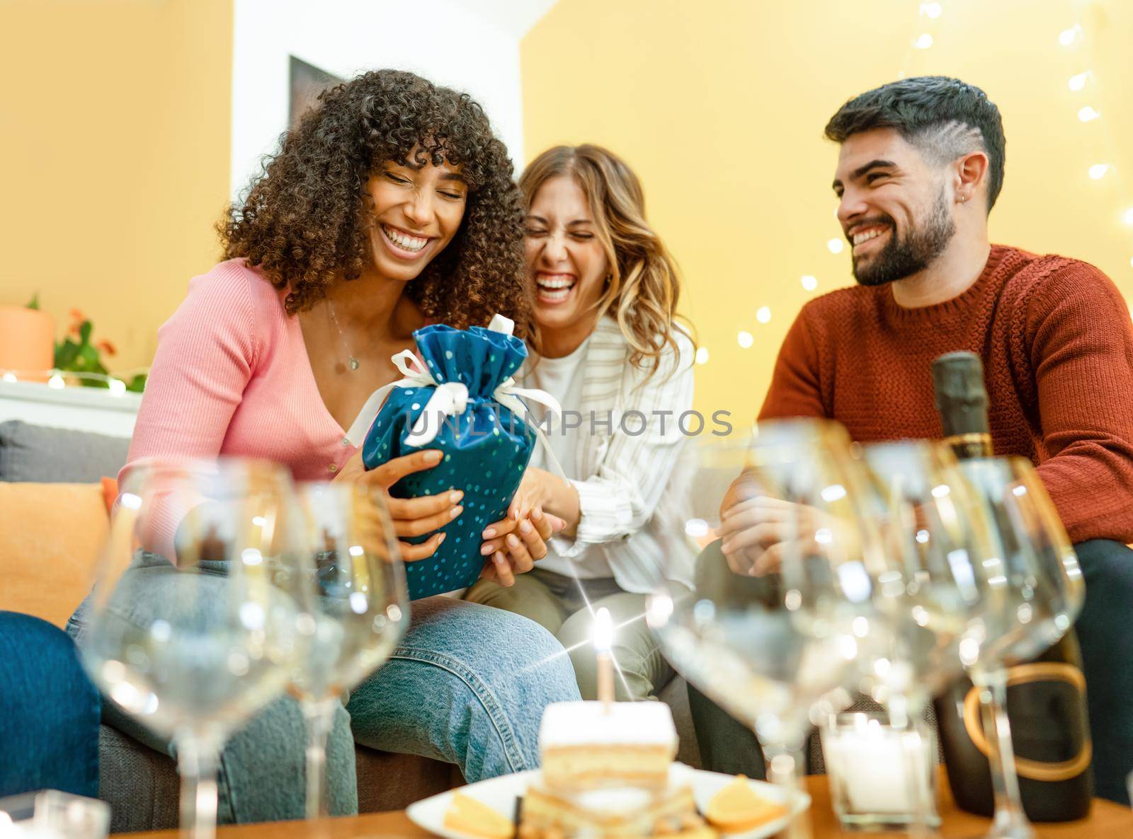 Happy birthday moment with multiracial real people at home laughing celebrating with champagne, cake with candle light effect. Amused black Hispanic young woman receives a gift from her best friends by robbyfontanesi