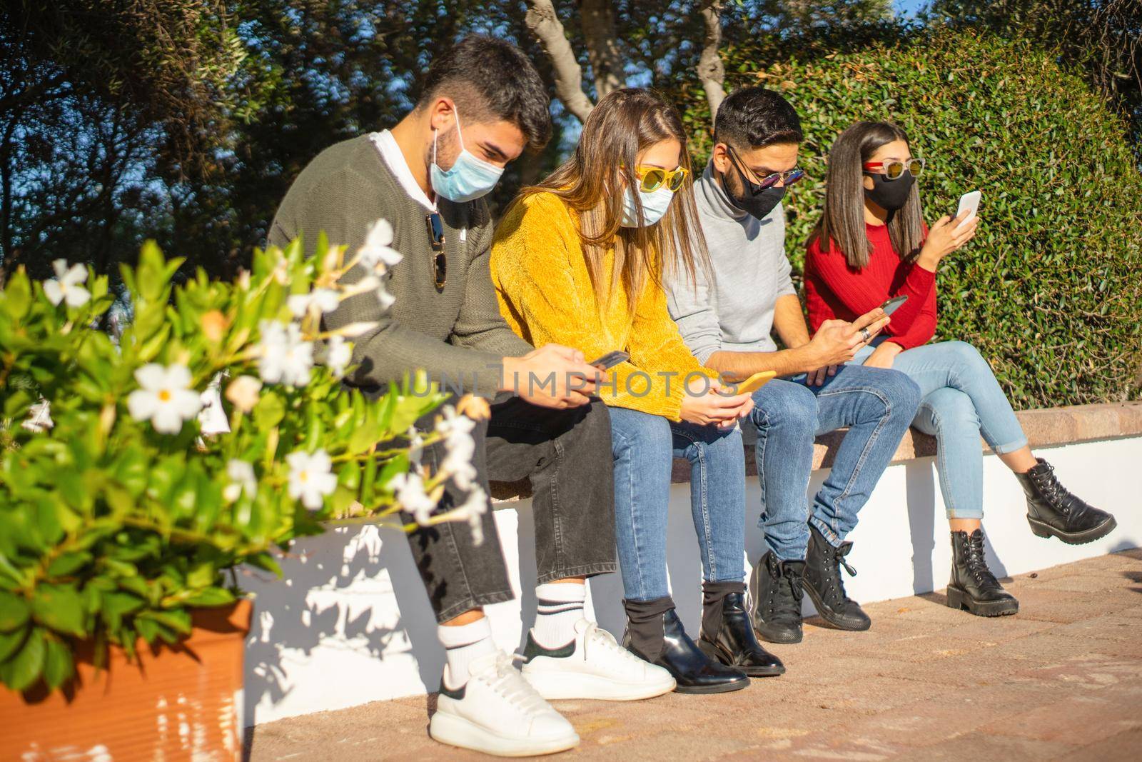 New normal social distancing for people in urban activities: group of millennial sitting on a city wall with protective mask against Coronavirus pandemic using smartphone while wait outside to enter by robbyfontanesi