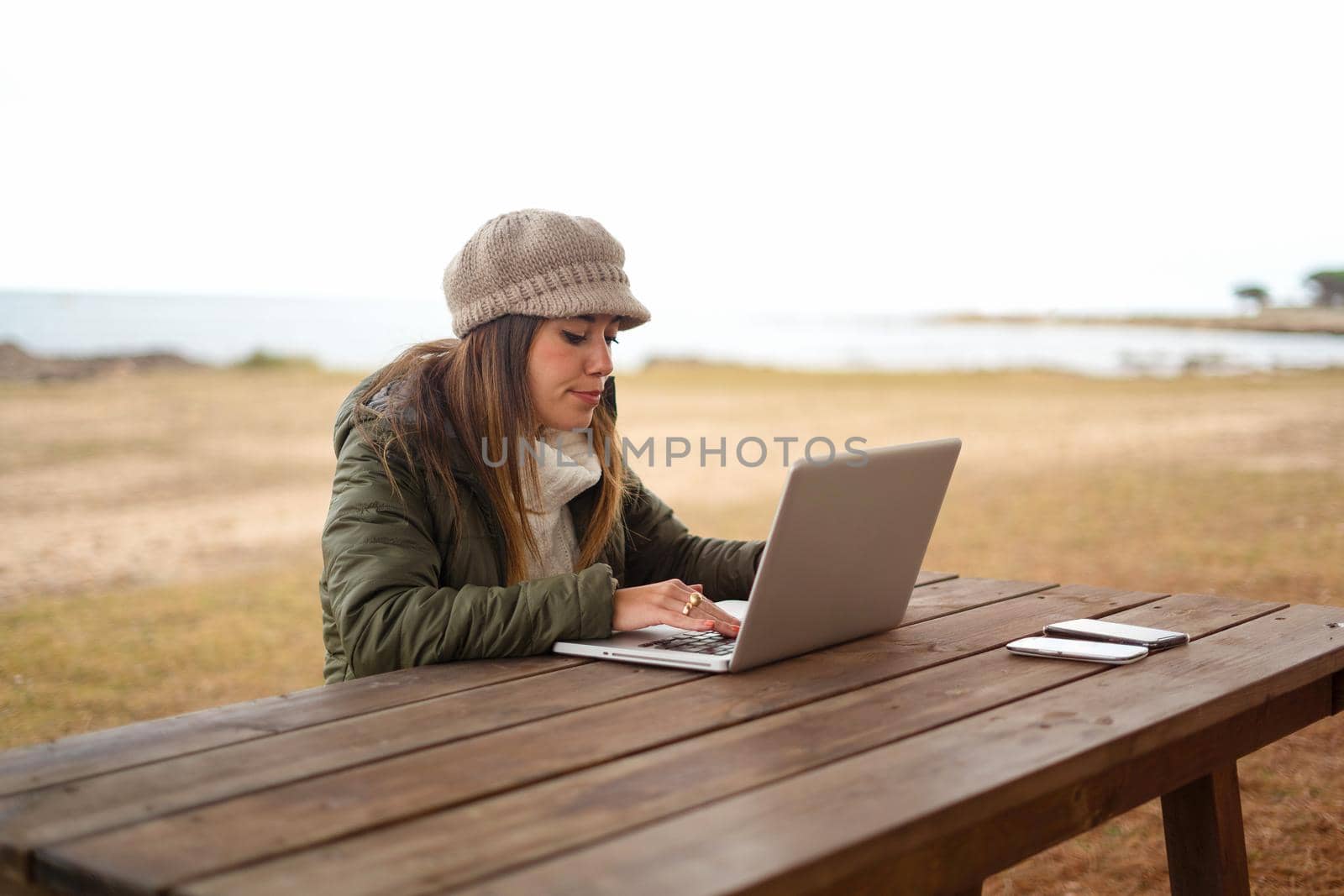 New normal job activities using internet connection outdoor: young Caucasian woman sitting at a wooden table in a sea park working at laptop managing her online business. University student in nature