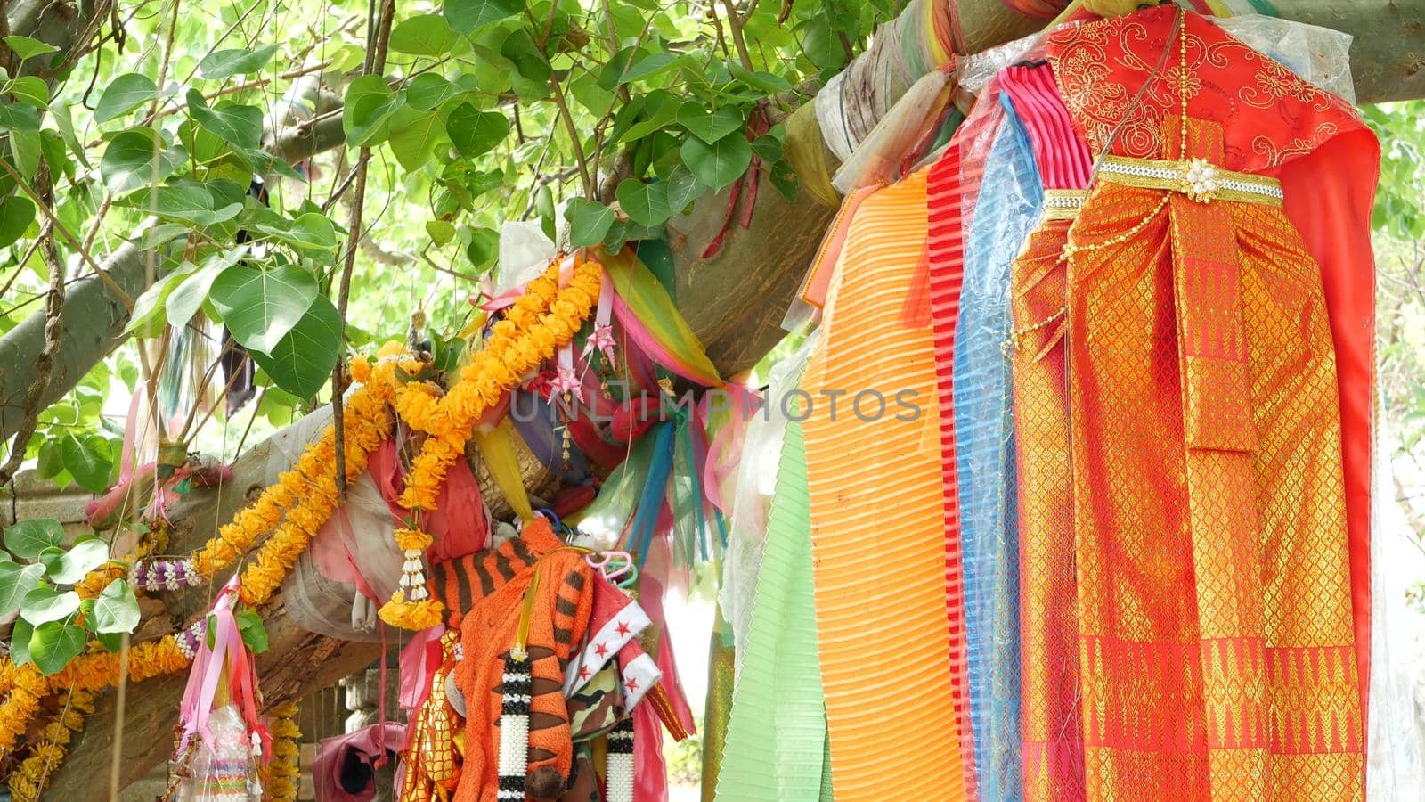Colorful offering clothes on tree near altar. Bunch of bright traditional clothes as gift to Thai spirits hanging on tree branches near animist altar.
