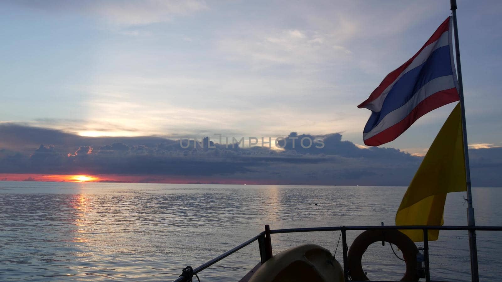 Boat floating near Thai flags in evening. Silhouette of anonymous person floating on boat on rippling sea near Thai and King of Thailand flags against cloudy sundown sky