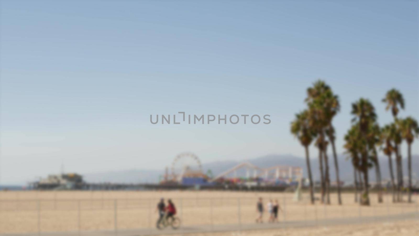 California beach aesthetic, people ride cycles on a bicycle path. Blurred, defocused background. Amusement park on pier and palms in Santa Monica american pacific ocean resort, Los Angeles CA USA.