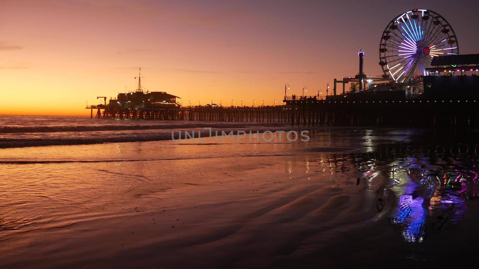 Twilight waves against classic illuminated ferris wheel, amusement park on pier in Santa Monica pacific ocean beach resort. Summertime iconic symbol of California glowing in dusk, Los Angeles, CA USA