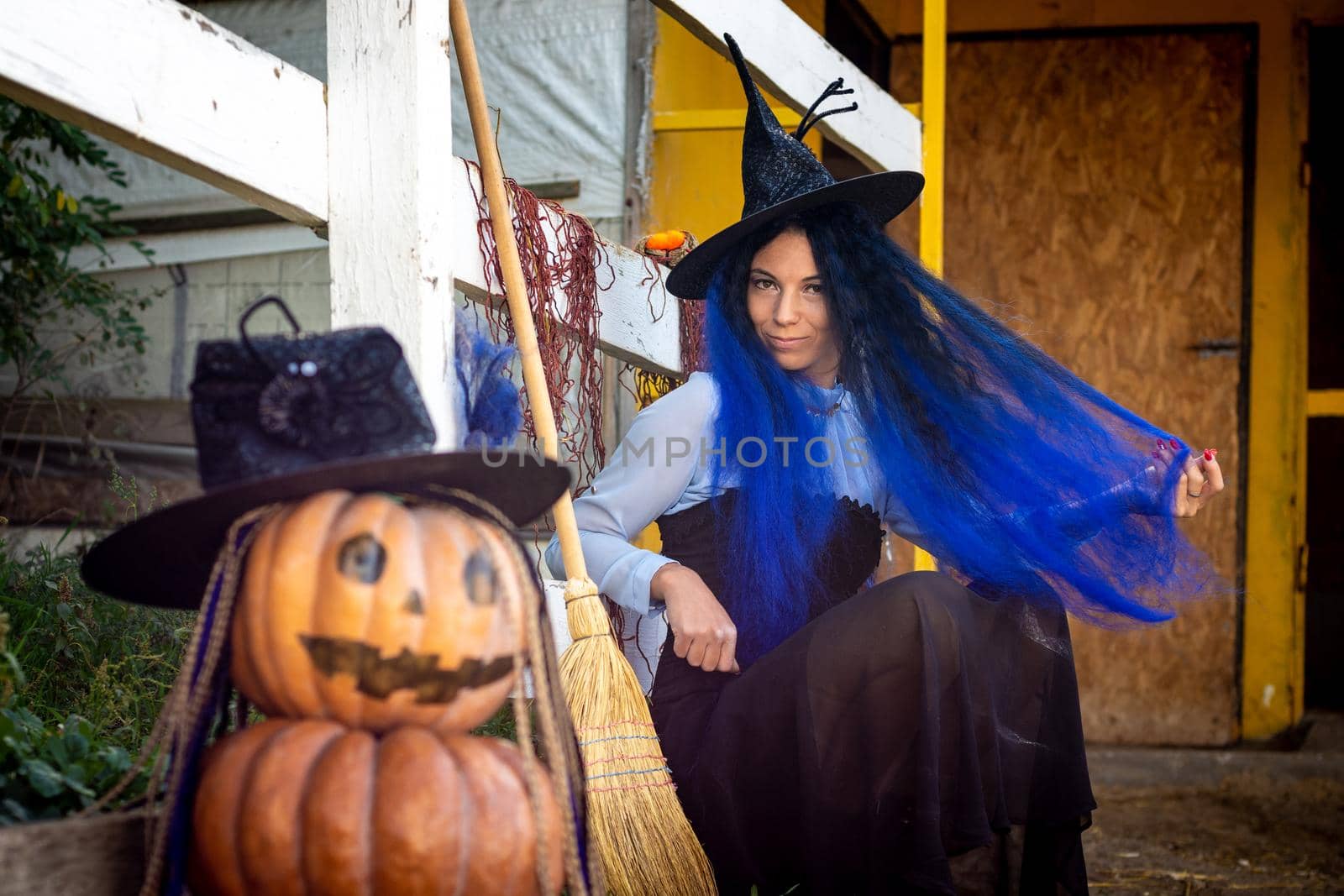 A witch with blue hair sits by the fence next to an evil pumpkin figure