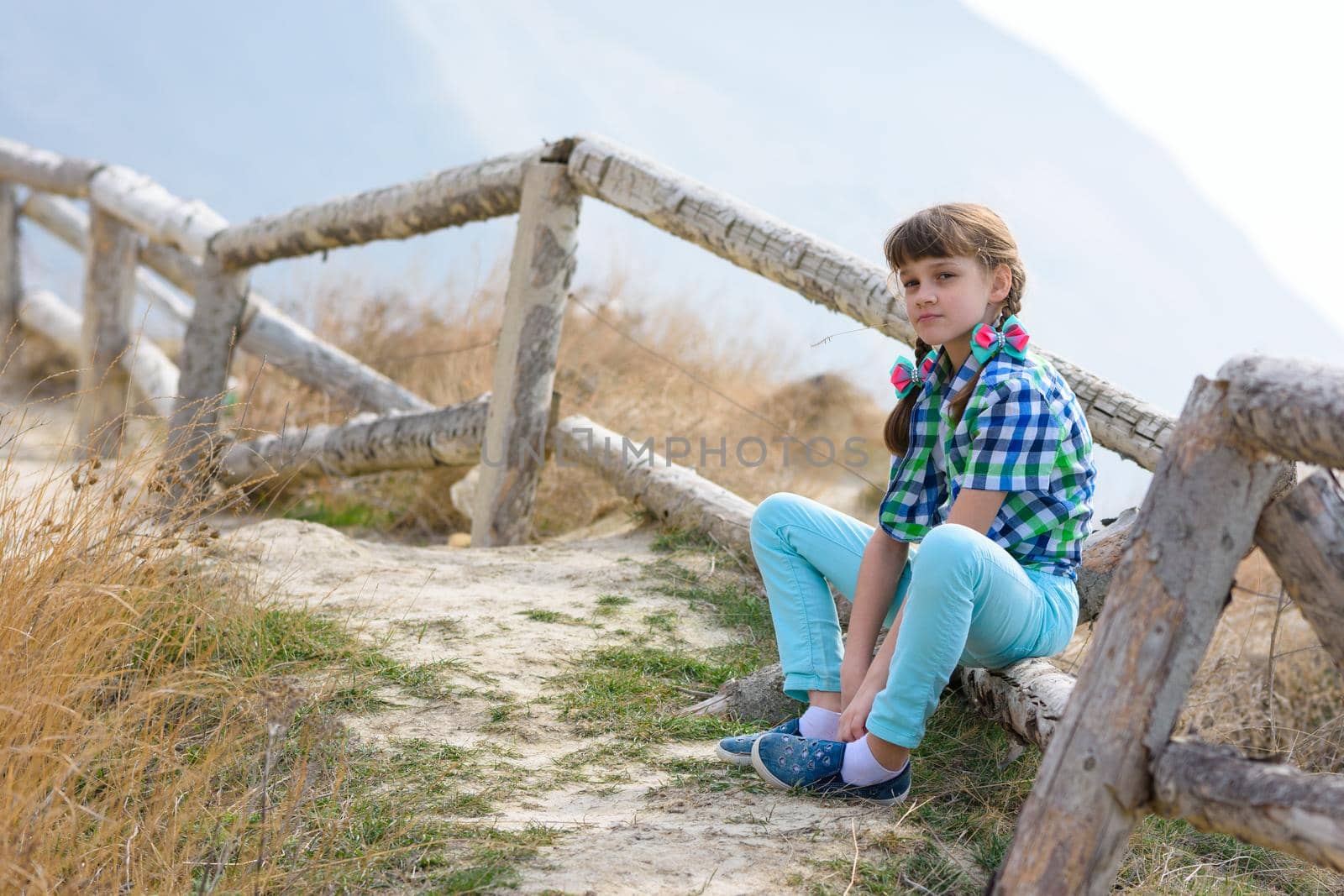 A girl sits on a fence made of a wooden blockhouse against the background of a landscape