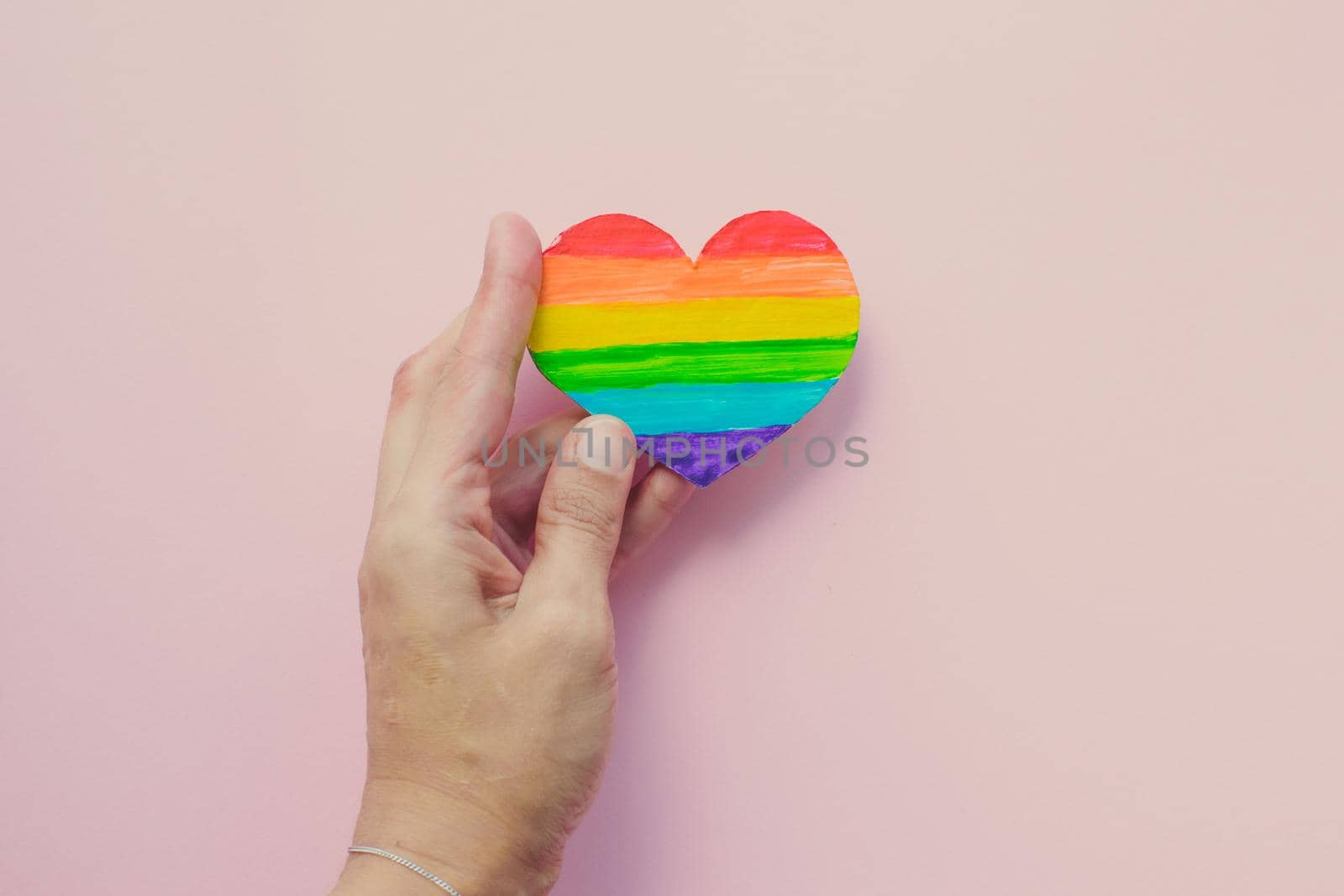 Female hands holding heart with rainbow stripes on pink background. LGBT pride flag, symbol of lesbian, gay, bisexual, transgender, social movements. Homosexual love, Human rights concept. Copy space.