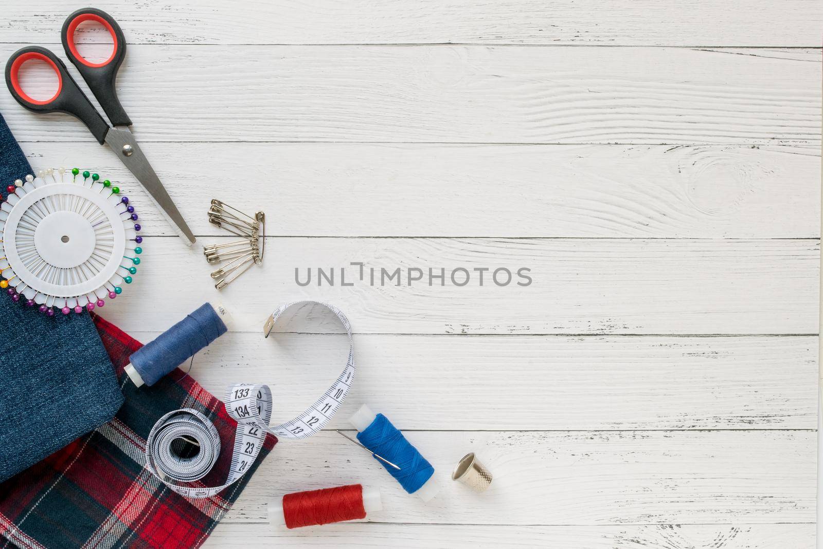 Sewing accessories, denim and plaid fabric on a white wooden background. Fabric, sewing threads, needle, pins, scissors, buttons and sewing centimeter. Copyspace