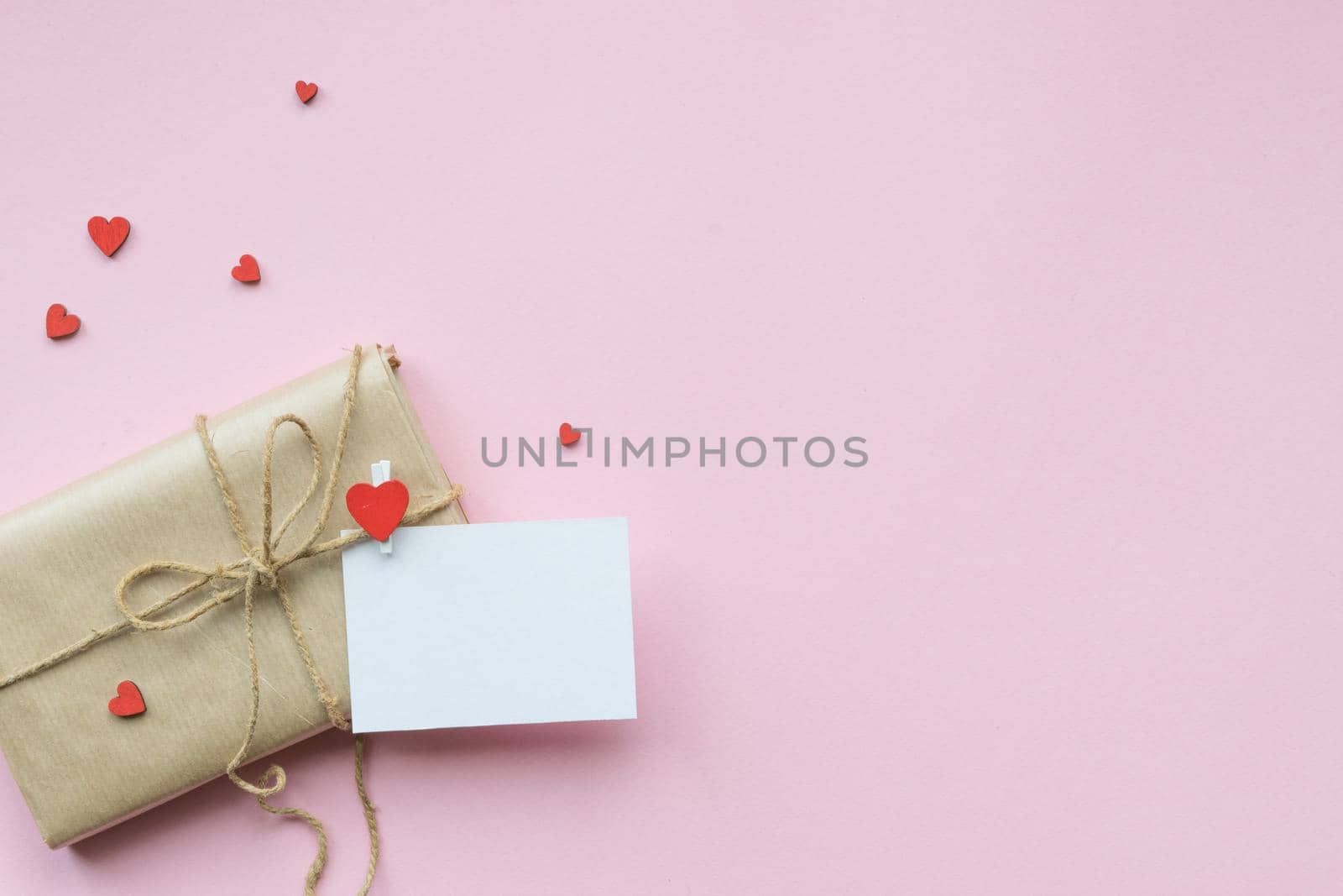Present wrapped in brown craft paper and tie on Light pink background. Gift with decorative red hearts and empty greeting card for Mothers Day, Wedding, Birthday and Valentines Day. Top view. Copyspace. Mockup