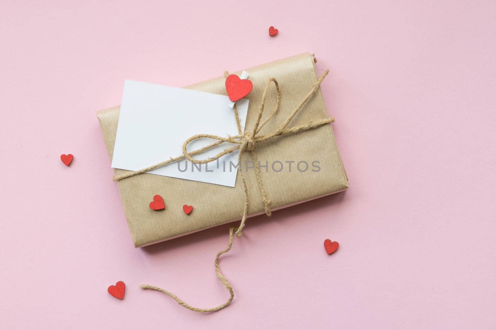 Present wrapped in brown craft paper and tie on Light pink background. Gift with decorative red hearts and empty greeting card for Mothers Day, Wedding, Birthday and Valentines Day. Top view. Copyspace. Mockup