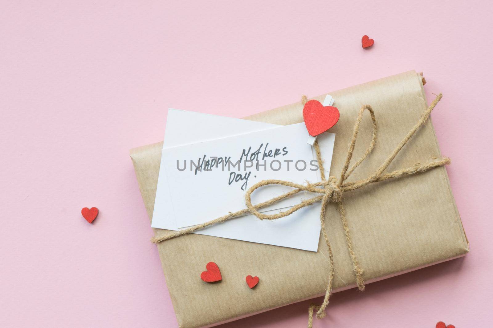 Present wrapped in brown craft paper and tie hemp string on Light pink background. Gift box with greetings on Mothers Day and decorative red hearts. Top view. Copyspace