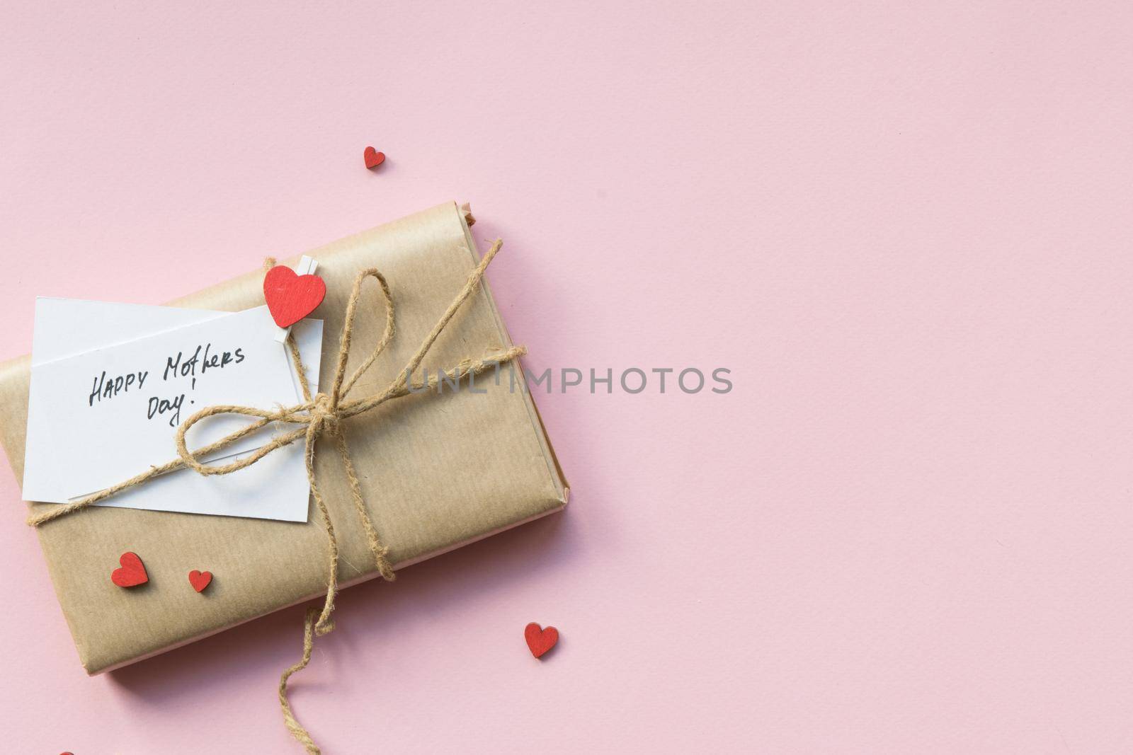 Present wrapped in brown craft paper and tie hemp string on Light pink background. Gift box with greetings on Mothers Day and decorative red hearts. Top view. Copyspace