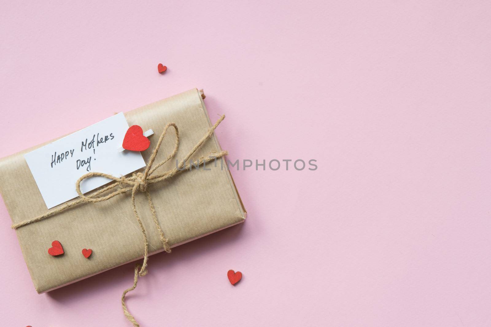 Presents wrapped in brown craft paper and tie hemp string on Light pink background. Gift box with greetings on Mothers Day and decorative red hearts. Top view. Copyspace