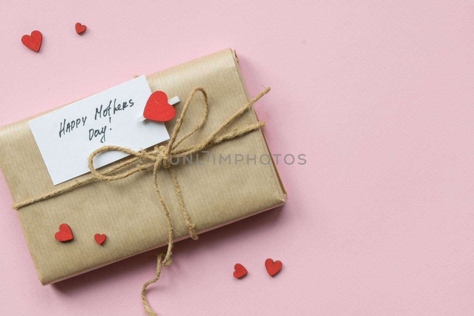 Present wrapped in brown craft paper and tie hemp string on Light pink background. Gift box with greetings on Mothers Day and decorative red hearts. Top view. Copyspace