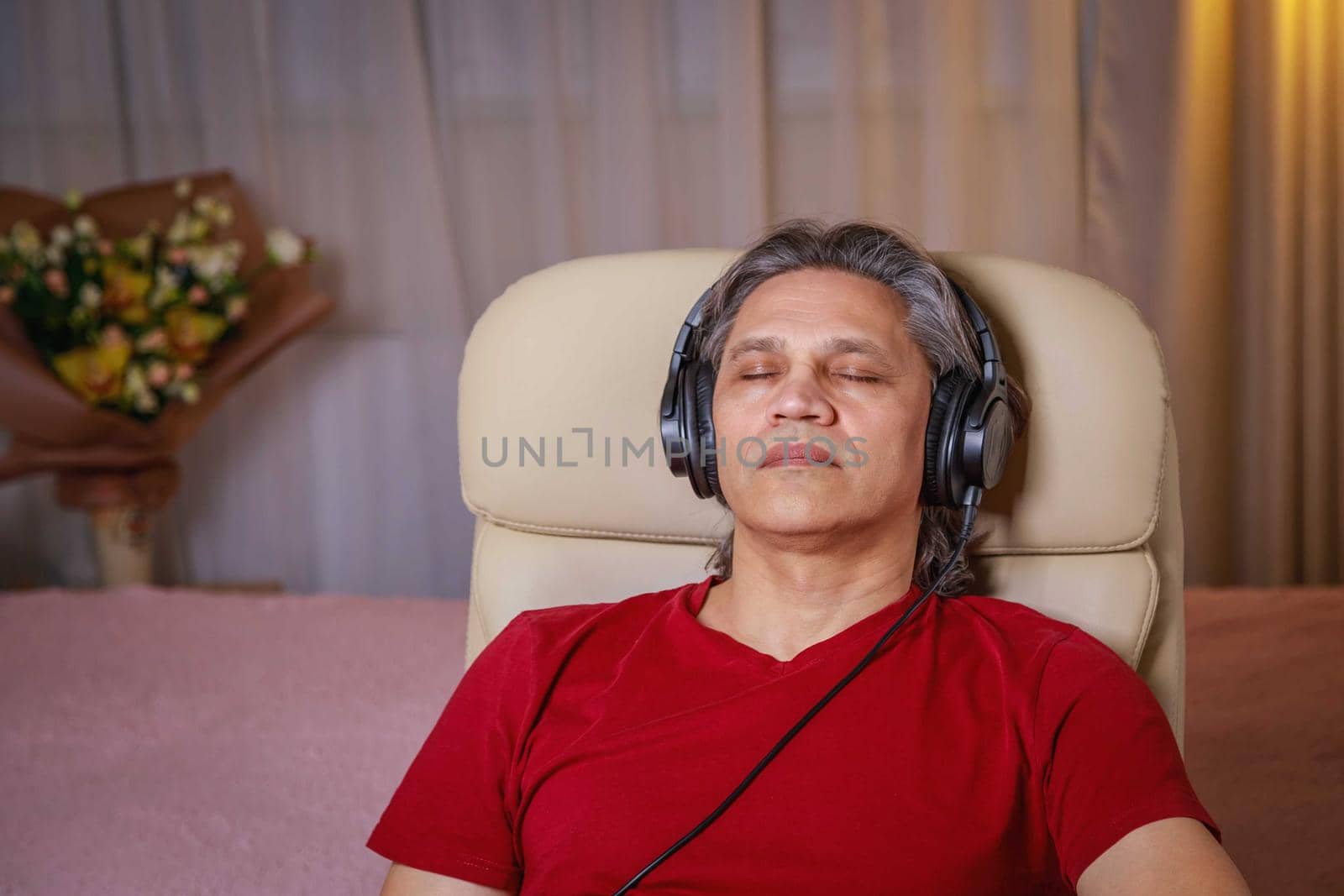 50-year-old man listens to music on headphones at home, sitting in a chair. by Yurich32