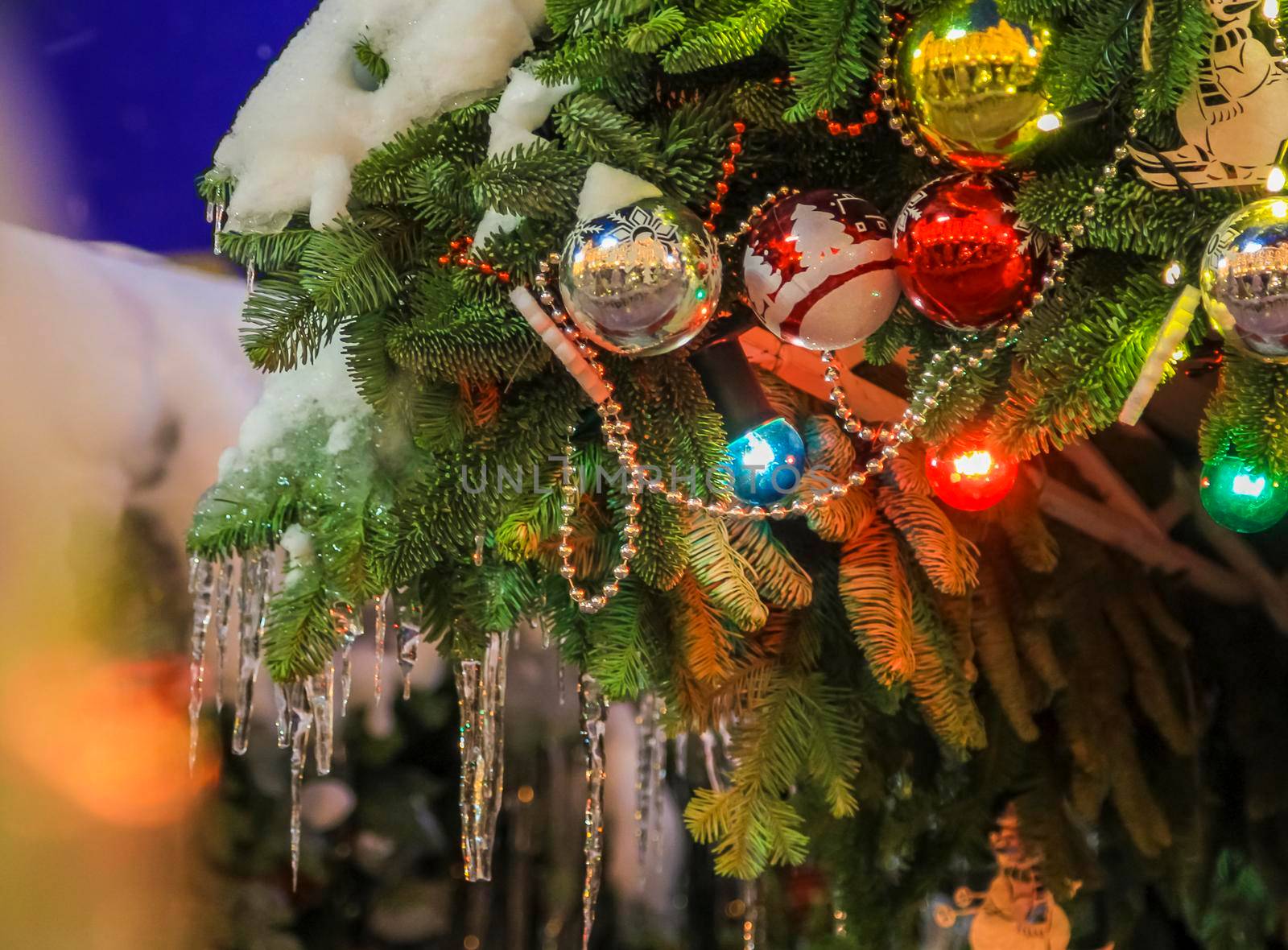 Branches of a green Christmas tree with a Christmas toy. Snow lies on the branches and icicles hang down. Bright garland.