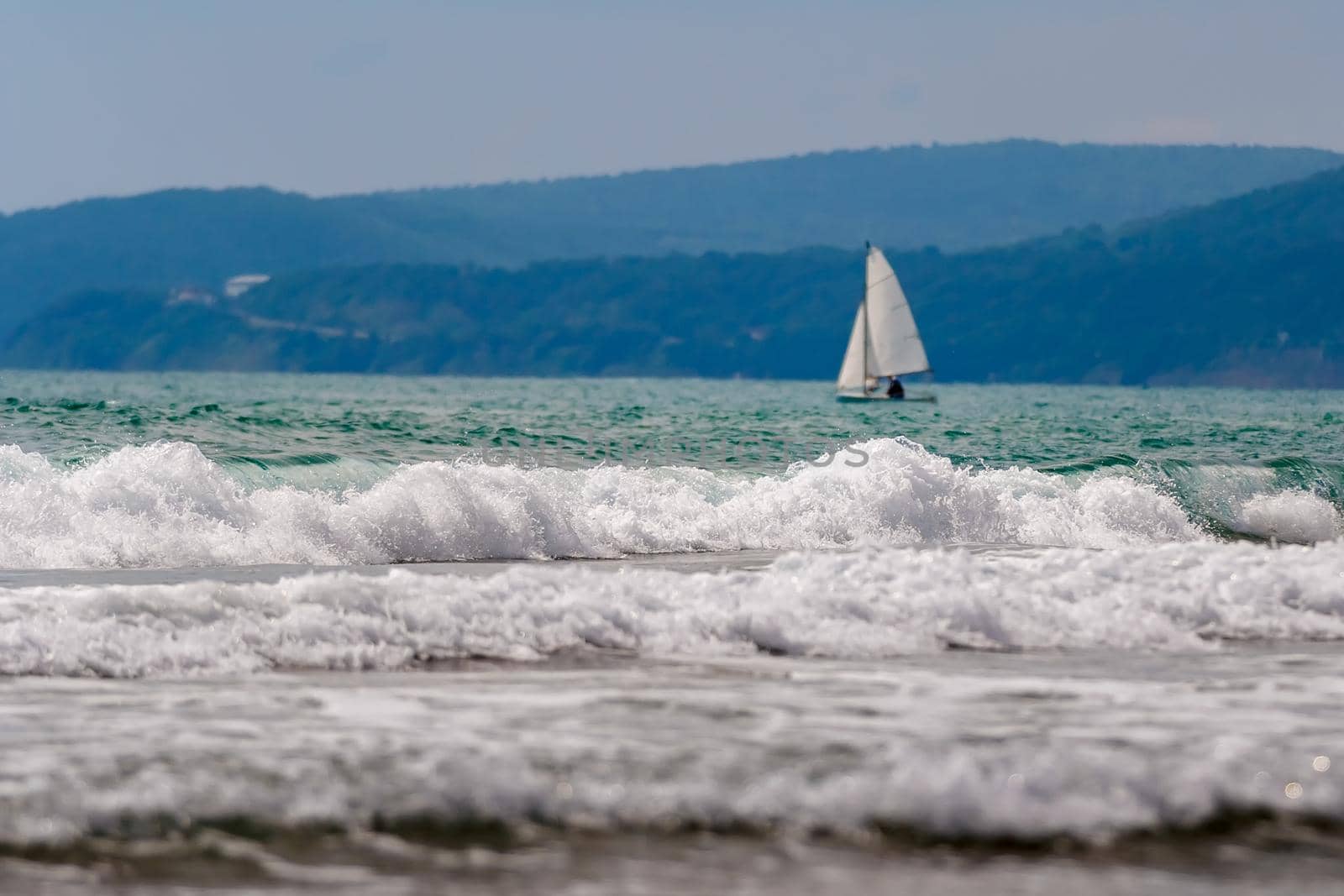 Seascape, small waves with white foam at the top against the background of blue water. A lone sailboat can be seen in the distance. A great place to relax and reflect