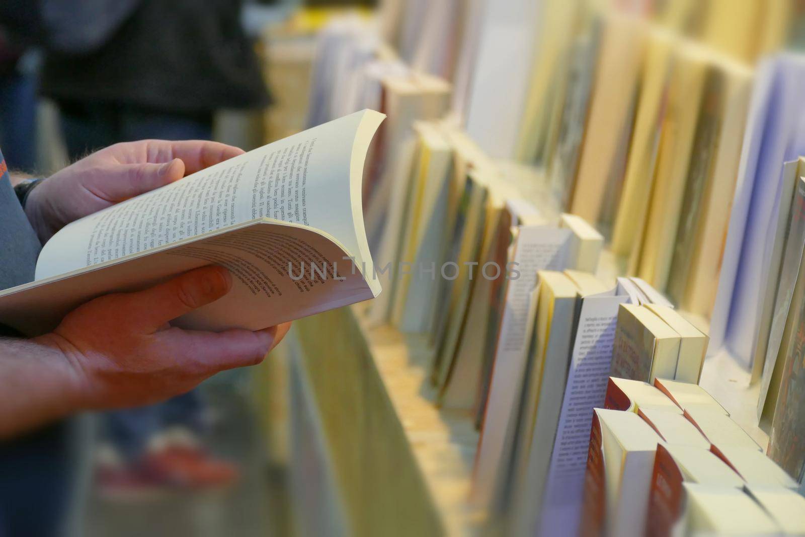 Close up perspective view of man hands holding an open book near book rows in book fair selective focus Turin Italy May 10 2019 by lemar