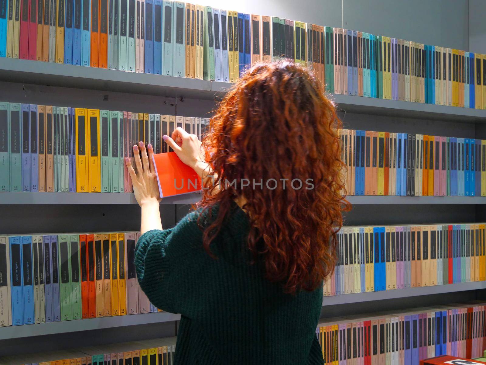 Female red haired teenager choosing a book from colorful rows in a bookstore