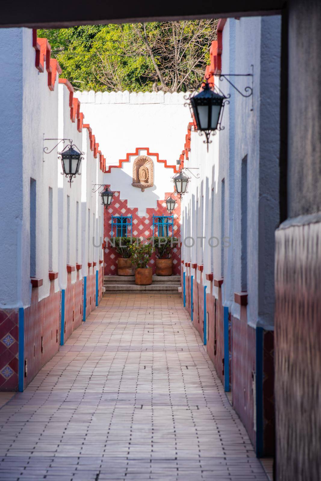 Hall in Mexico City with white walls, classic hanging lamps, and plants at the end. Long corridor with brown tiled floor and classic Hispanic architecture. Colorful Mexican neighborhood
