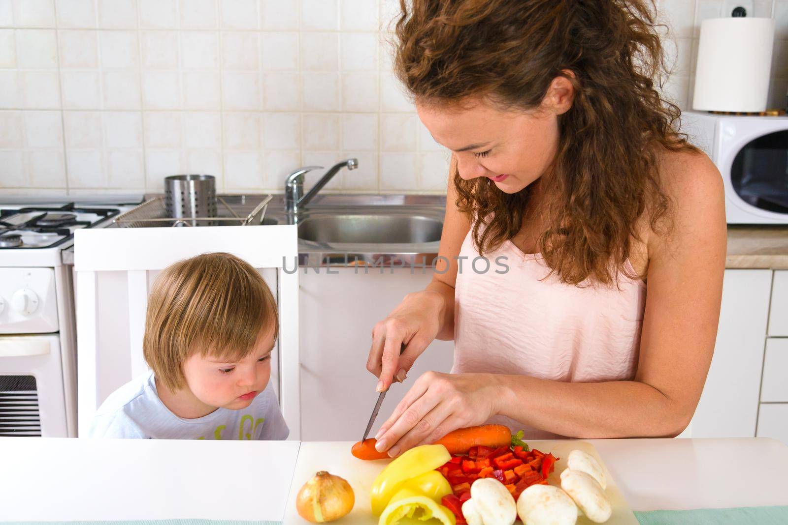 Young women preparing healthy launch from vegetables in white kitchen while his son toddler with Down Syndrome curiously looking. In white kitchen.