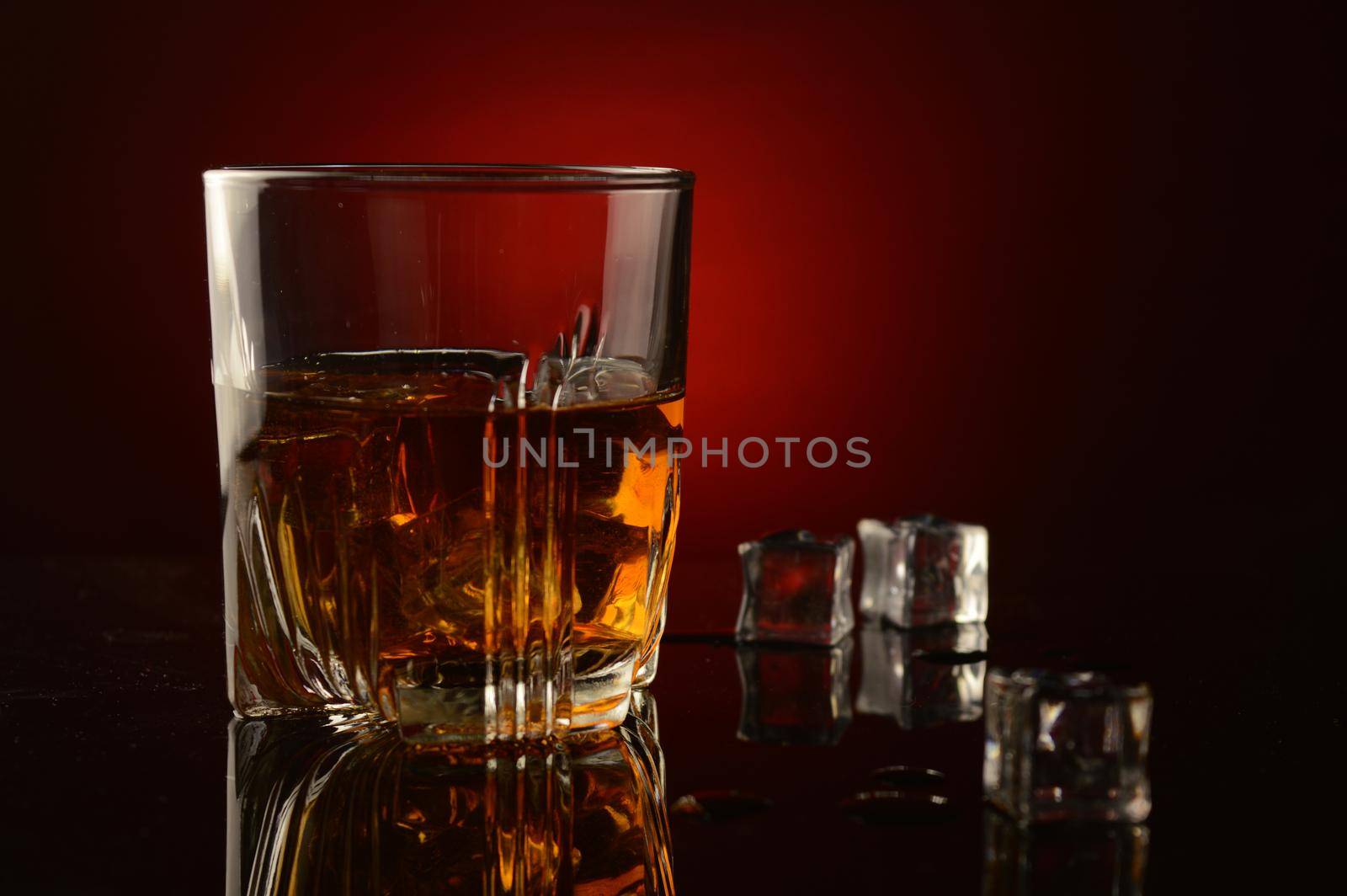 A closeup of a glass full of whiskey and ice over a dark red background with a shallow depth of field on the glass.