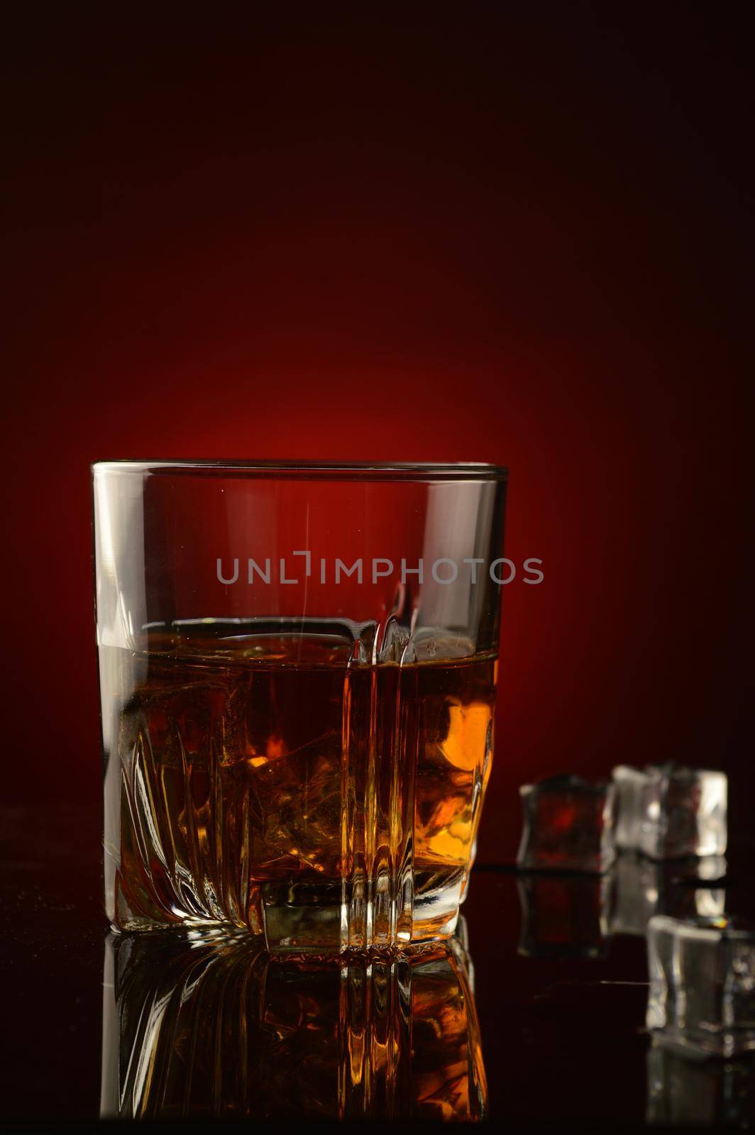 A closeup of a glass full of whiskey and ice over a dark red background with a shallow depth of field on the glass.