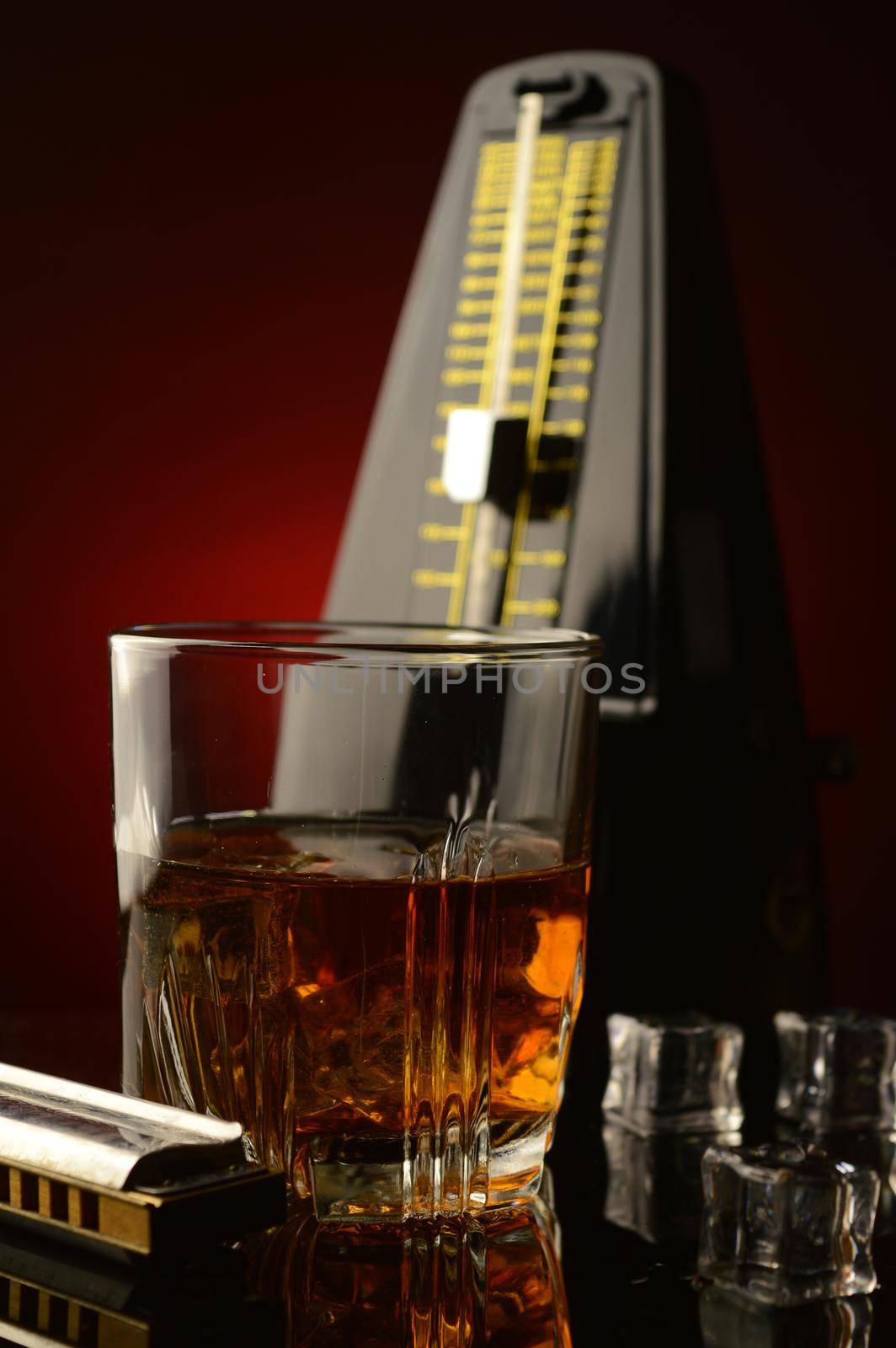A concept of rythem and blues utilizing a glass of whiskey with a harmonica and a musical metronome over a reflective dark red and black background.