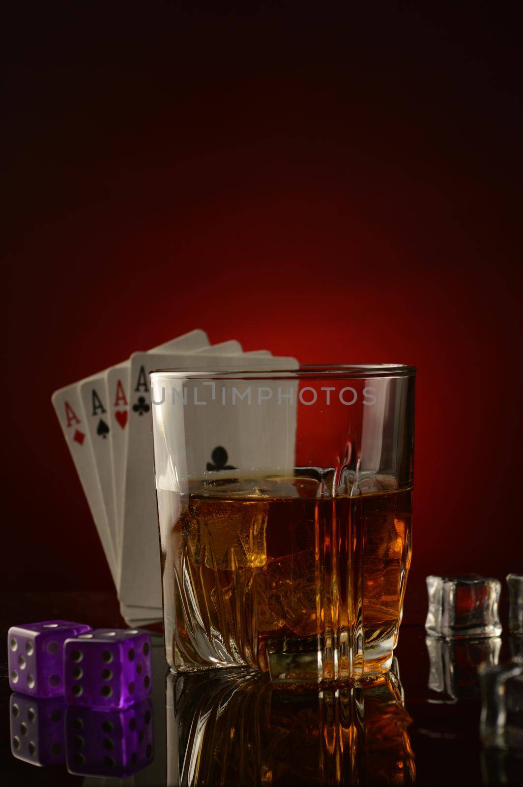 A conceptual image of a gambling mans drink focused on a glass of whiskey with four aces and pair of casino dice over a dark atmospheric background.