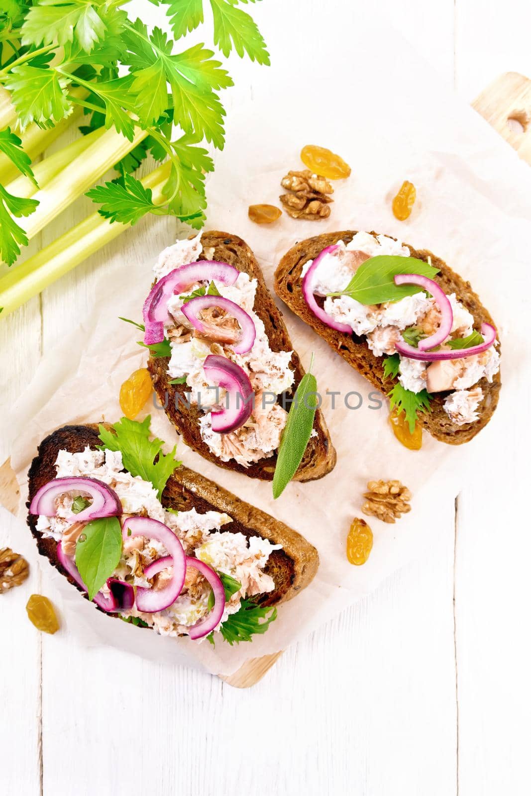 Salmon, petiole celery, raisins, walnuts, red onions and curd cheese salad on toasted bread with green lettuce on paper on a light wooden board background from above