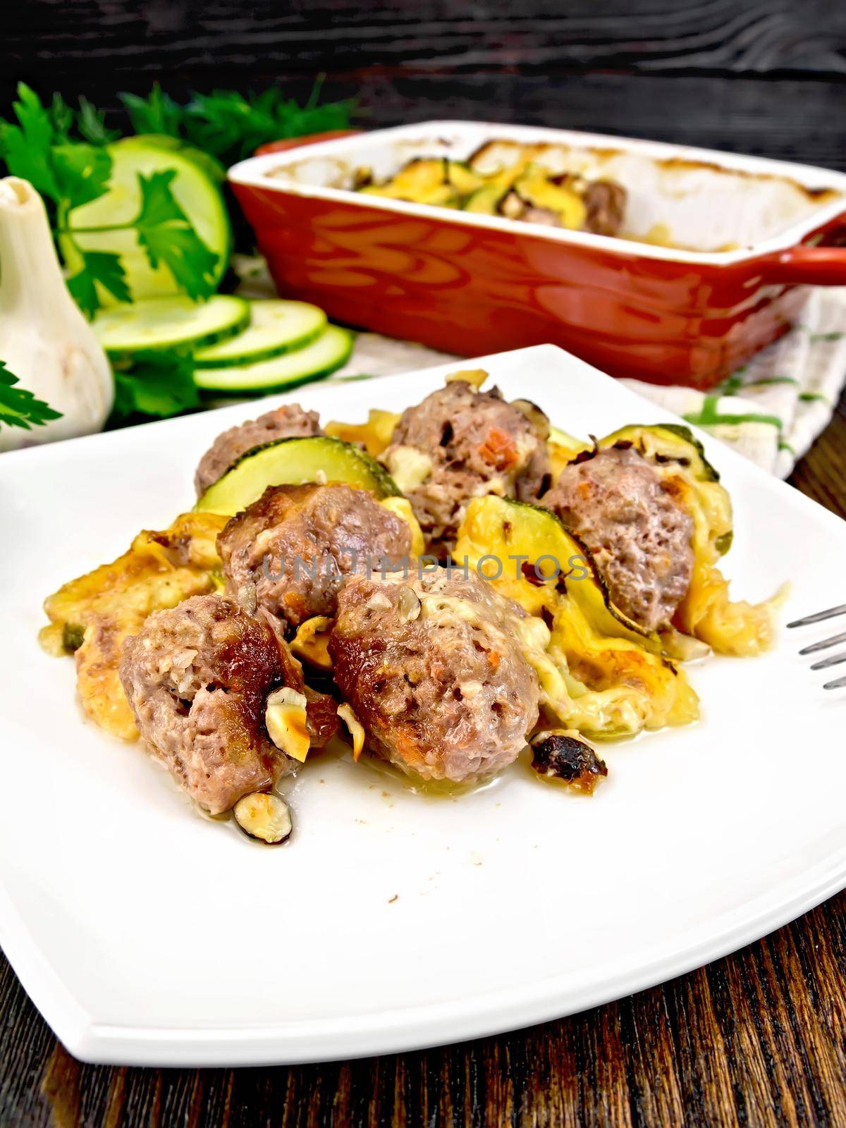 Meatballs baked with zucchini, cheese and nuts in a plate, napkin, garlic, parsley and fork on a wooden plank background