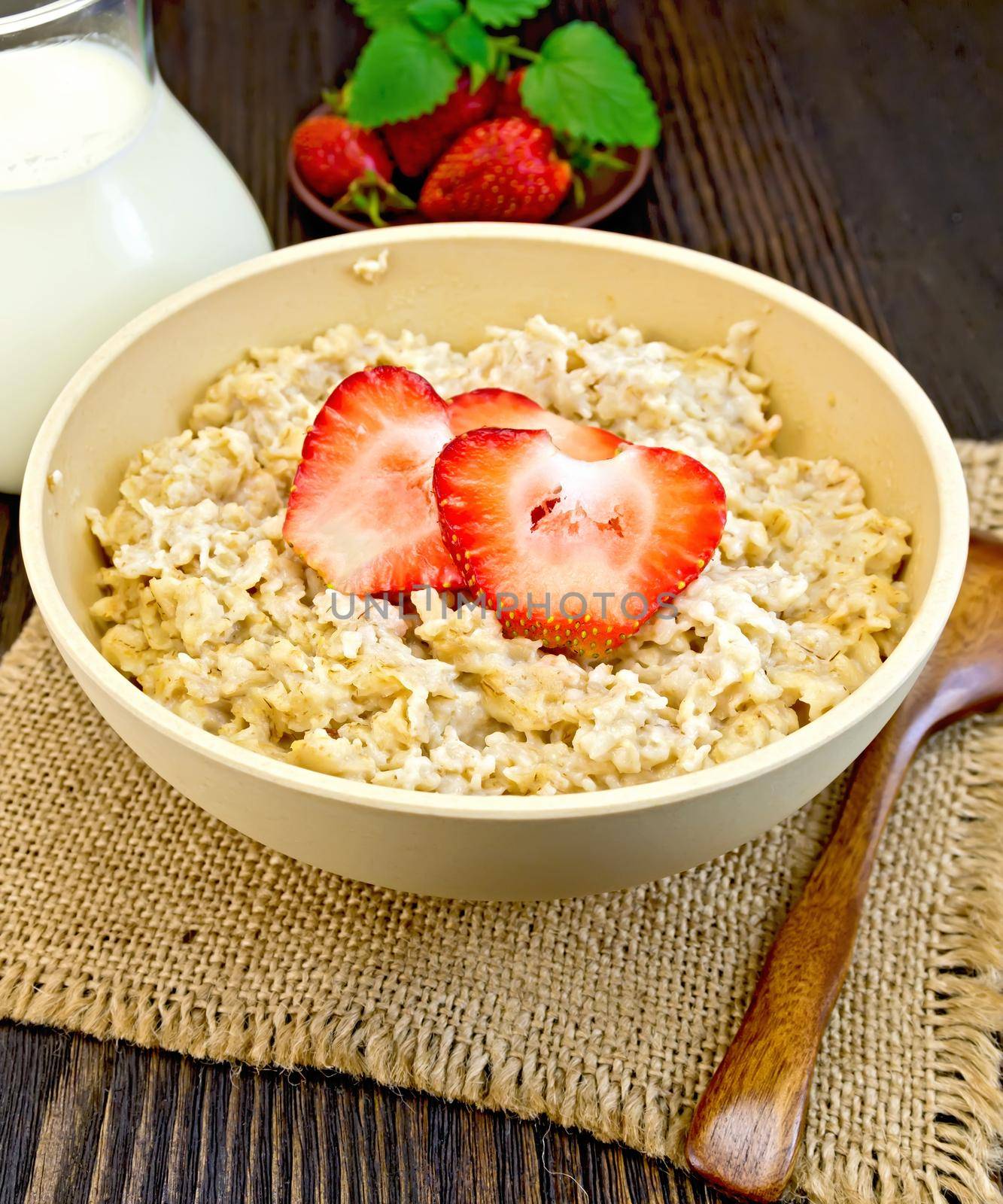 Oatmeal in a bamboo bowl with strawberries on a napkin of burlap, spoon, milk in a glass jar on the background of dark board