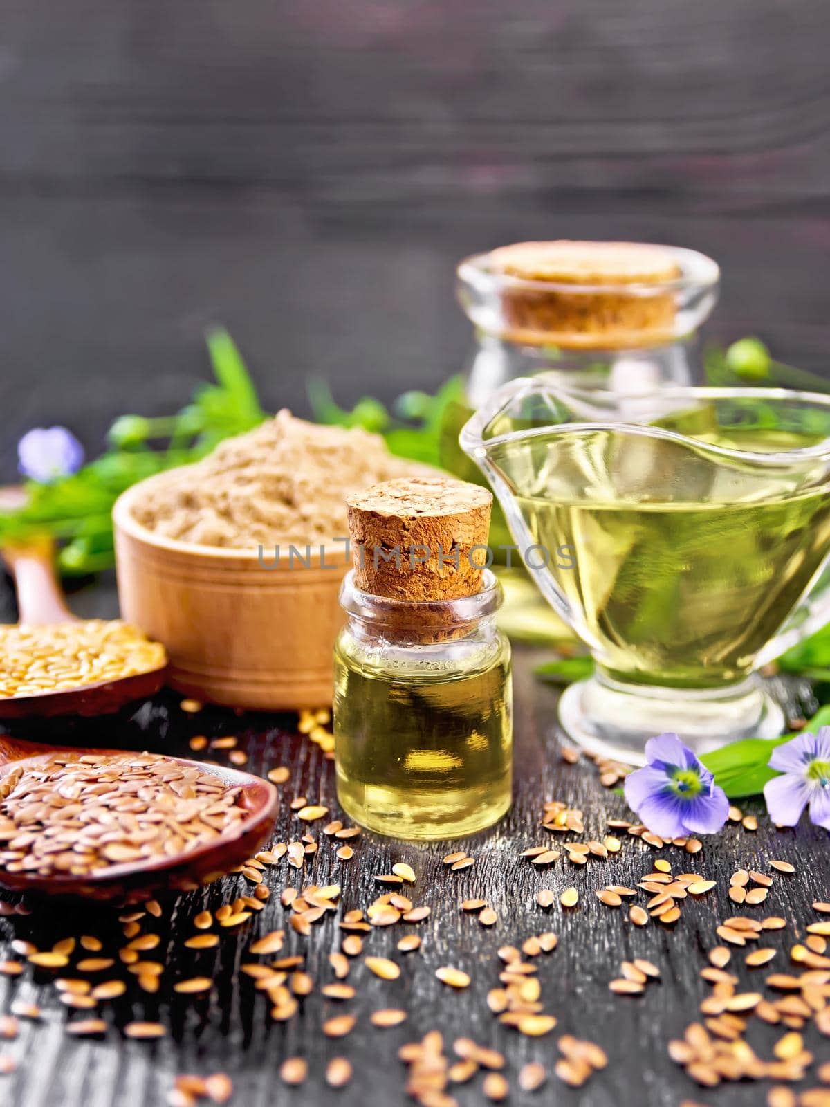 Linseed oil in two glass jars and a sauce boat with white and brown flax seeds in spoons, flour in a bowl, leaves and flowers on wooden board background