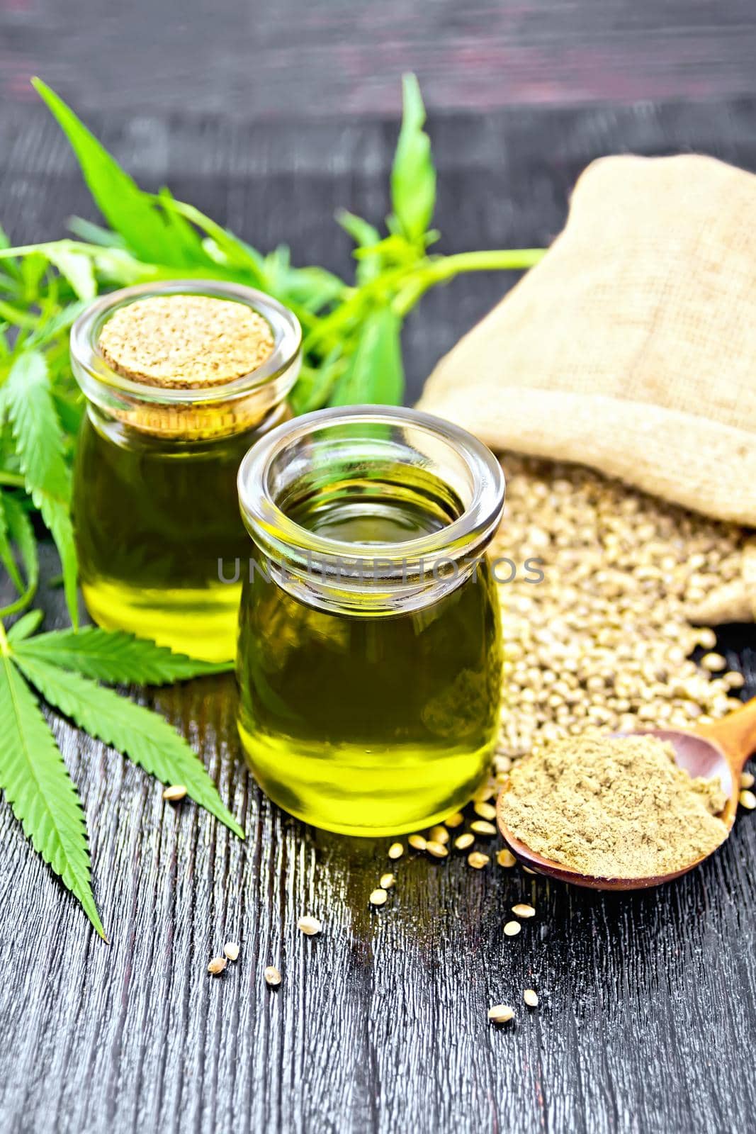Hemp oil in two glass jars, grain in a bag and on the table, flour in a spoon, leaves and stalks of cannabis on a wooden board background