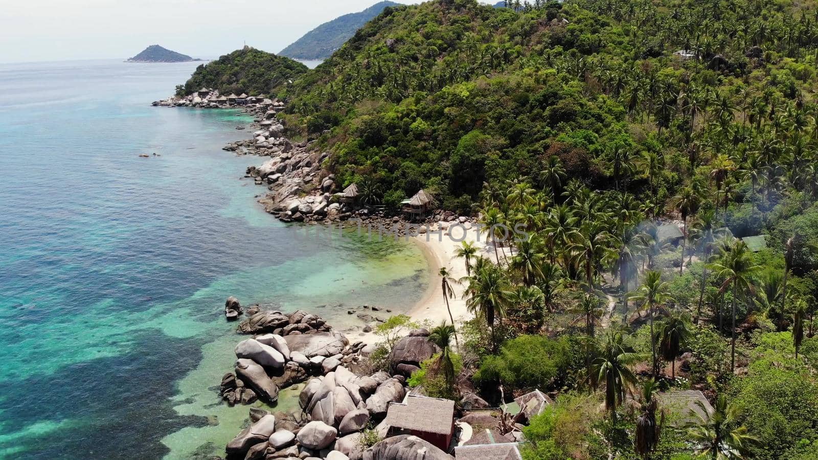 Bungalows and green coconut palms on tropical beach. Cottages on sandy shore of diving and snorkeling resort on Koh Tao paradise island near calm blue sea on sunny day in Thailand. Drone view